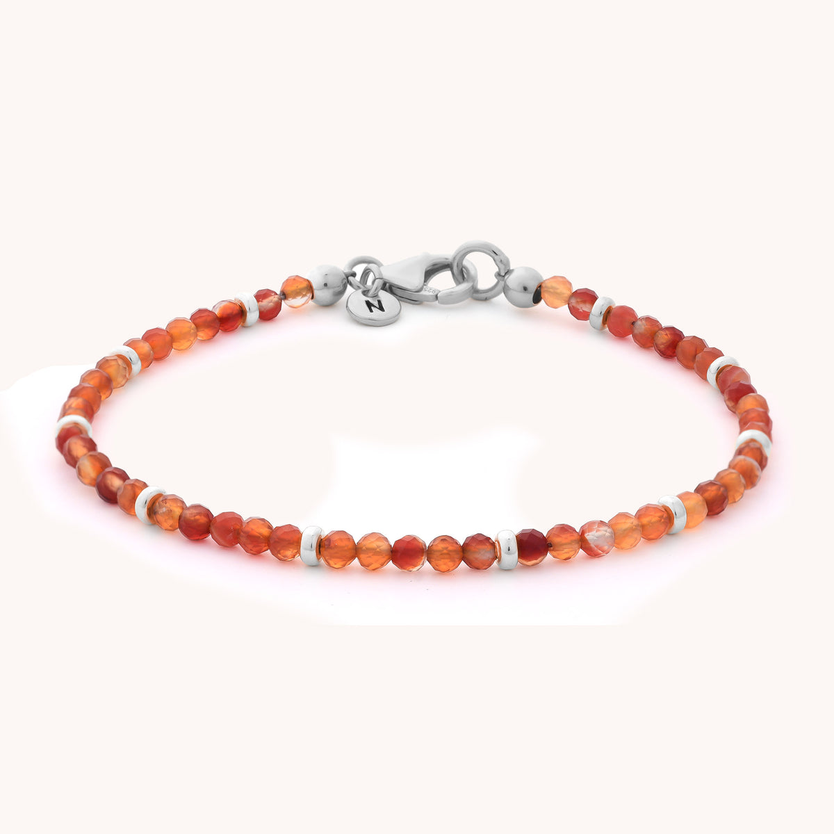 Introducing the Carnelian Orbit Bracelet - July: A delicate, handcrafted jewelry piece adorned with small, faceted orange carnelian beads and accented by silver spacers. Finished with a silver clasp and a petite round charm engraved with the letter N, this bracelet is beautifully showcased against a light background.