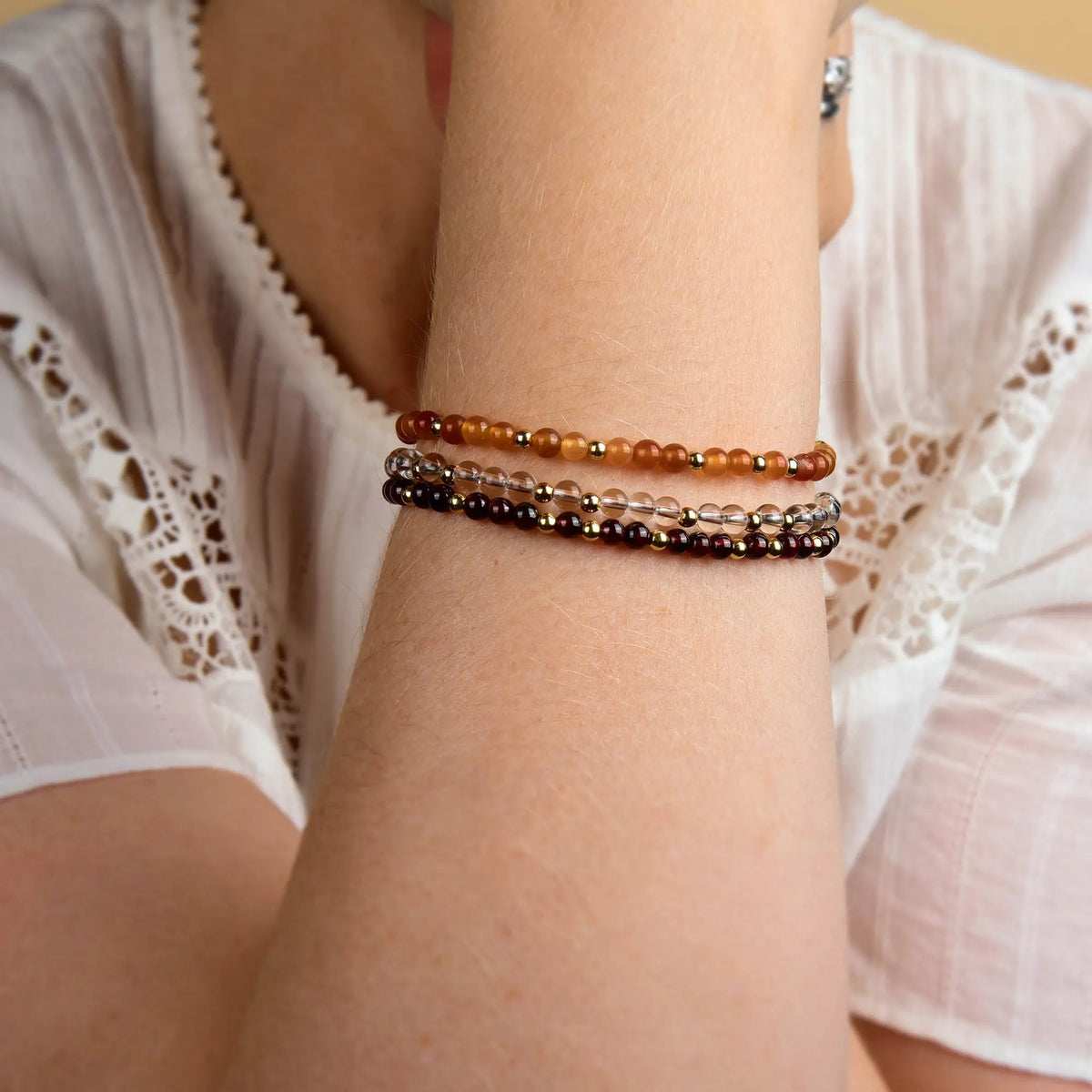 A person wearing a white blouse rests their arm on their chin, displaying three beaded bracelets in amber, clear, and dark brown tones on their wrist, including the Garnet Jewel Bead Birthstone Bracelet—featuring natural garnet and sterling silver beads—as a stylish tribute to the January birthstone.