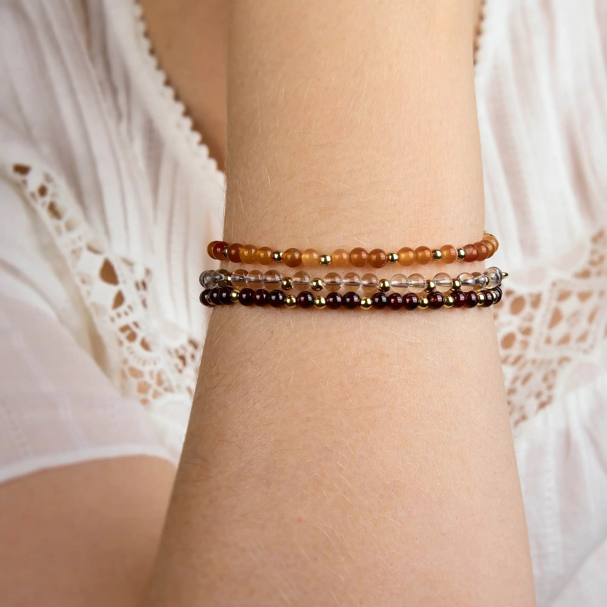 A person wearing a white lace sleeve displays three beautifully handcrafted Jewel Bead Birthstone Bracelets—one red, one clear, and one brown—on their forearm. Each artisan accessory is made in the UK and can be worn as a meaningful birthstone bracelet or styled simply as an elegant Jewel Bead Bracelet.