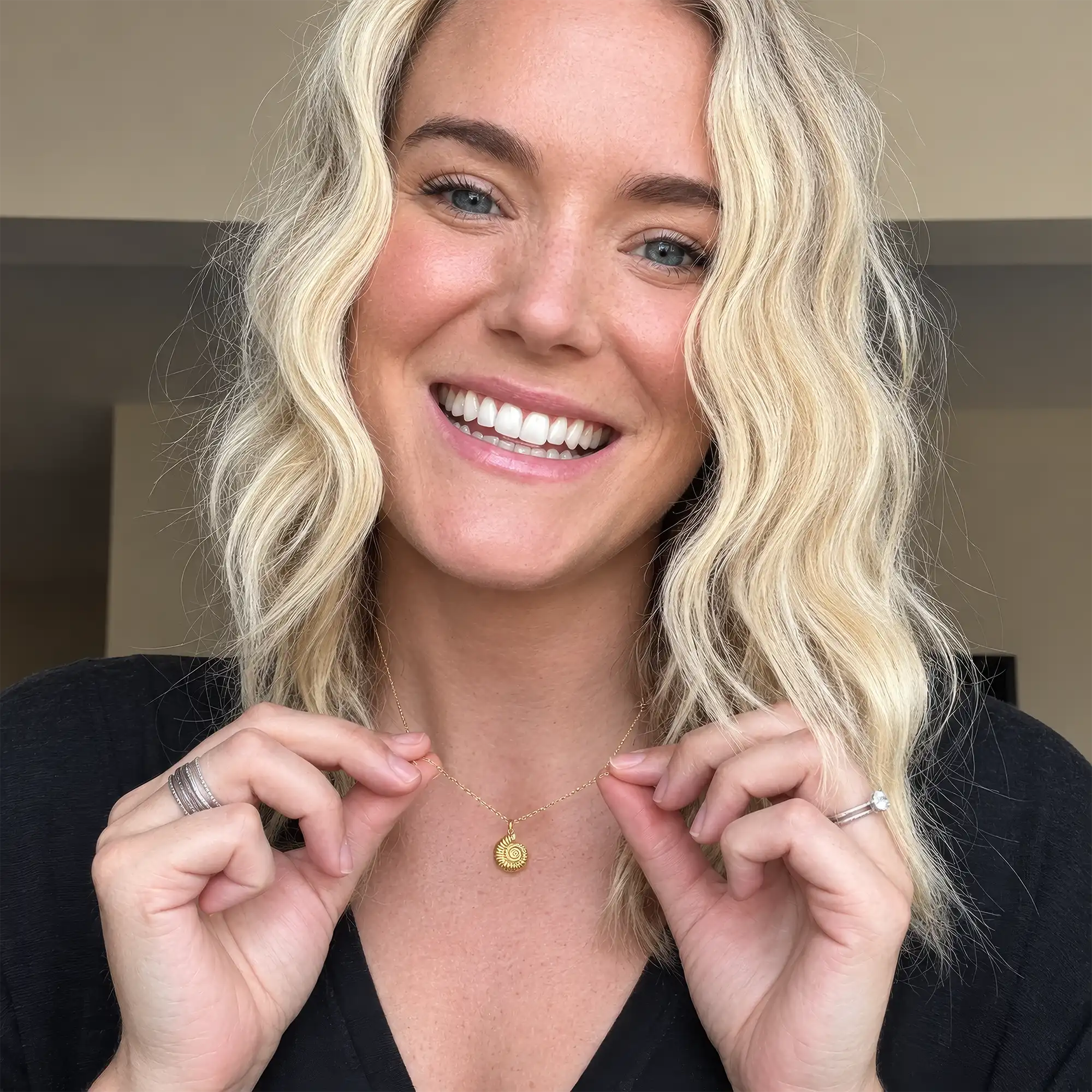 A smiling person with wavy blonde hair, dressed in a black top, holds up the Chloe Dixon | Ammonite Necklace—an elegant piece featuring a spiral pendant crafted from gold plated sterling silver—and displays rings on both hands.