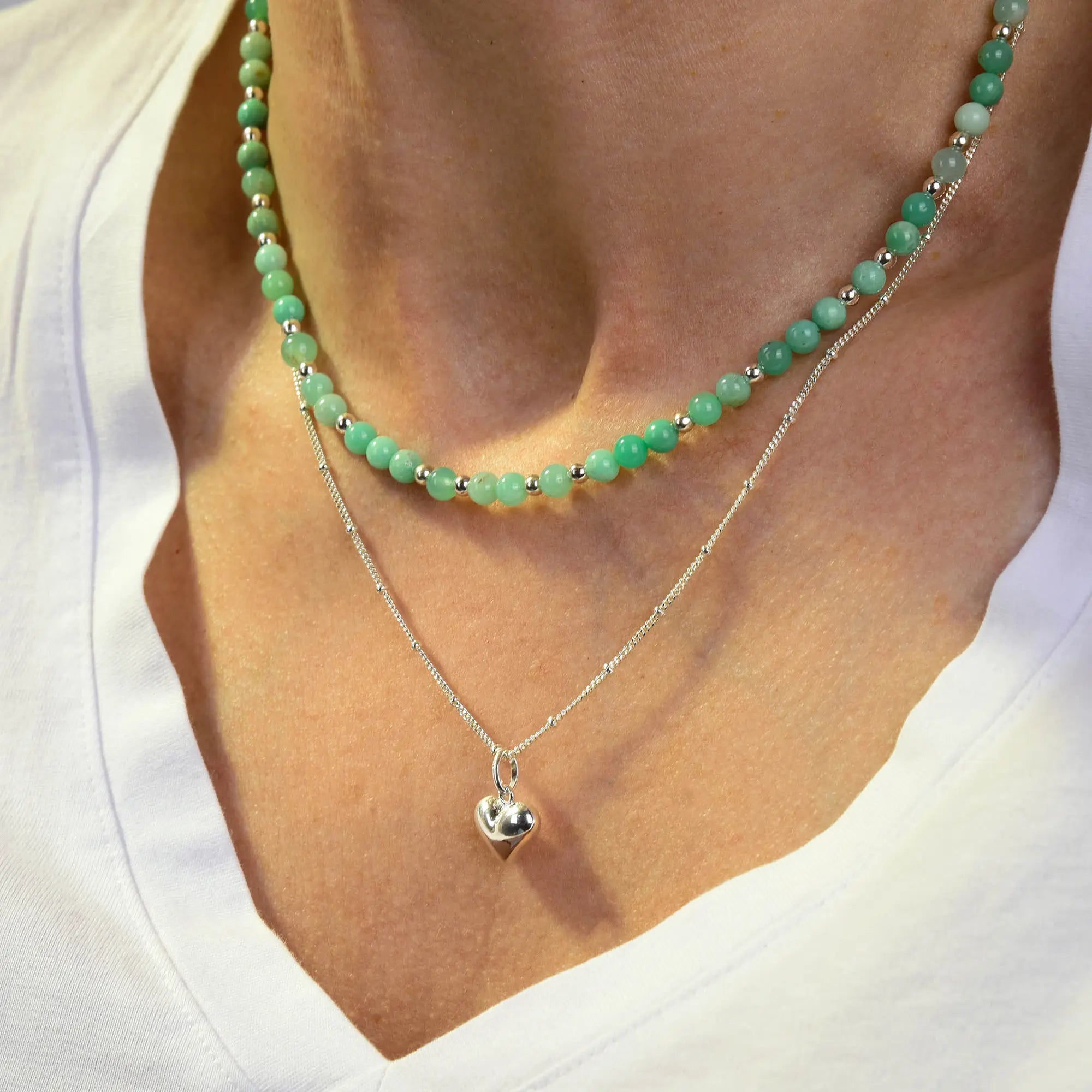 A person wearing a white V-neck shirt with two necklaces: one is an eco-friendly sterling silver chain adorned with a shiny heart-shaped pendant, and the other is the Jewel Bead Birthstone Necklace, featuring vibrant turquoise gemstone beads.