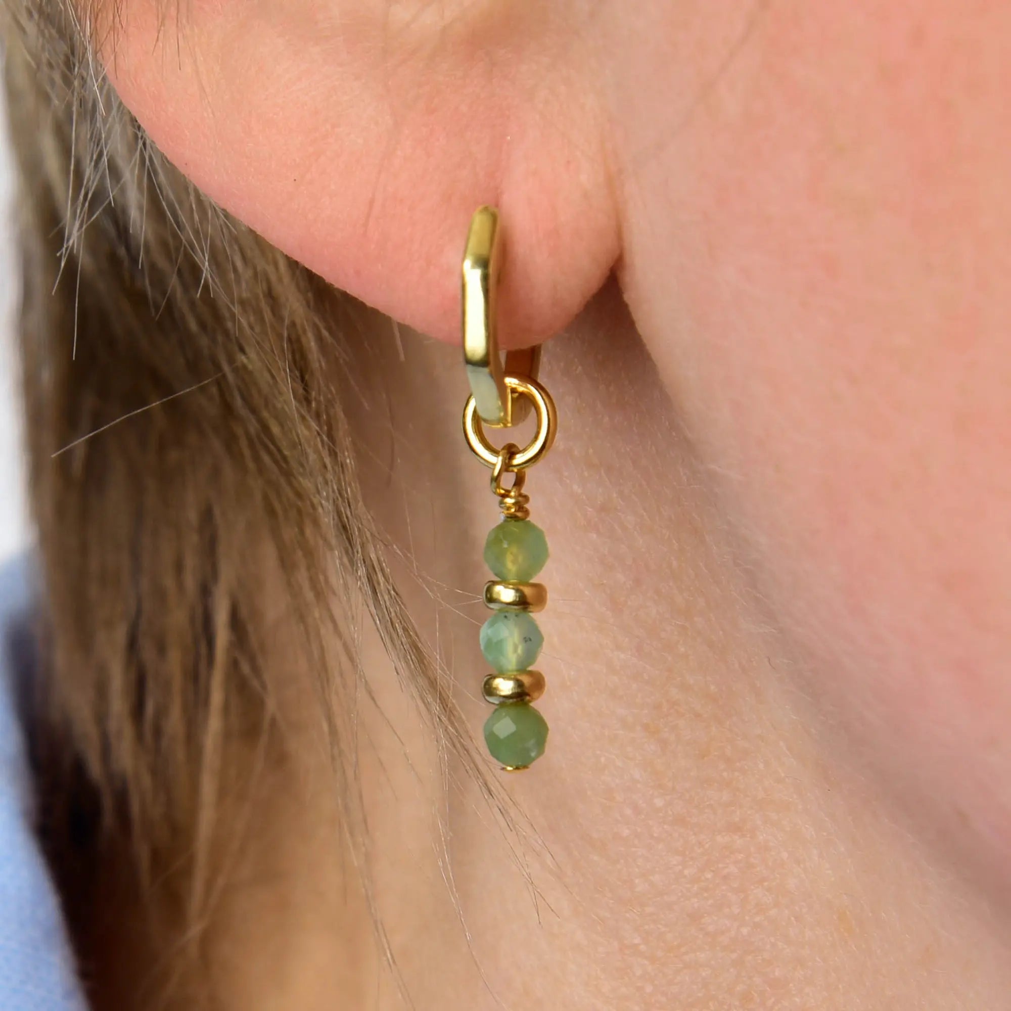A close-up of the Chrysoprase Linear Huggie Birthstone Earrings (May) shows a gold hoop adorned with three small green beads hanging vertically, elegantly highlighting the vibrant May birthstone on the wearer’s ear.
