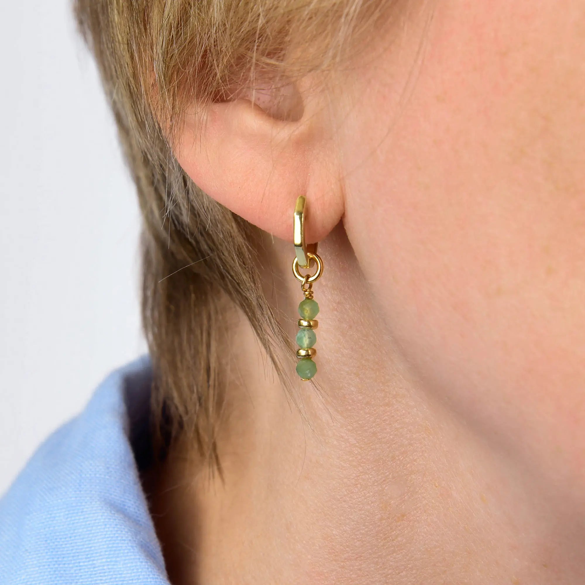 A close-up of a person’s ear wearing the Chrysoprase Linear Huggie Birthstone Earrings (May), featuring a gold hoop adorned with three small green beads hanging vertically for a perfect May birthstone accent. The person has short light brown hair and is dressed in a light blue shirt.