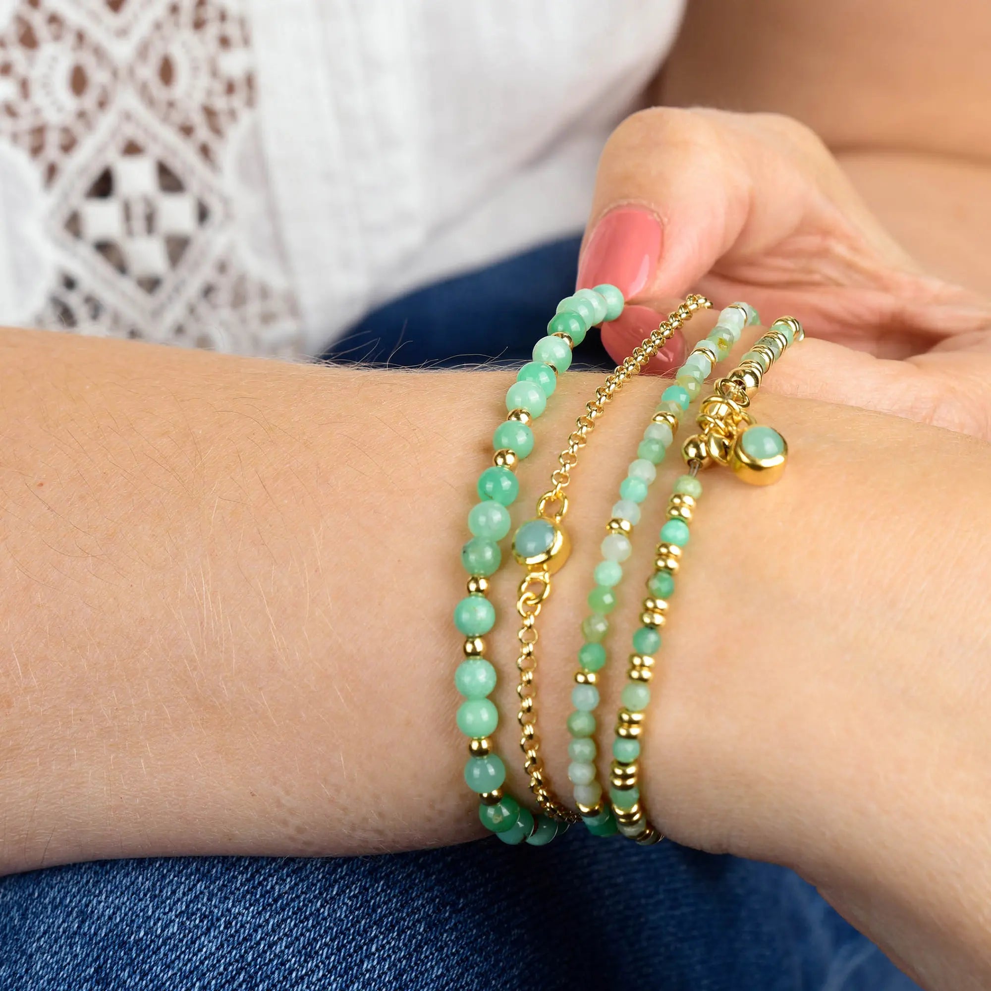 A person wearing a white lace top displays their wrist adorned with multiple green and gold beaded bracelets, including the Chrysoprase Birthstone Gem Dot Bracelet - May. Their nails are painted pink, making this look a perfect birthday gift for someone born in May or a stylish stack featuring a gold plated bracelet.