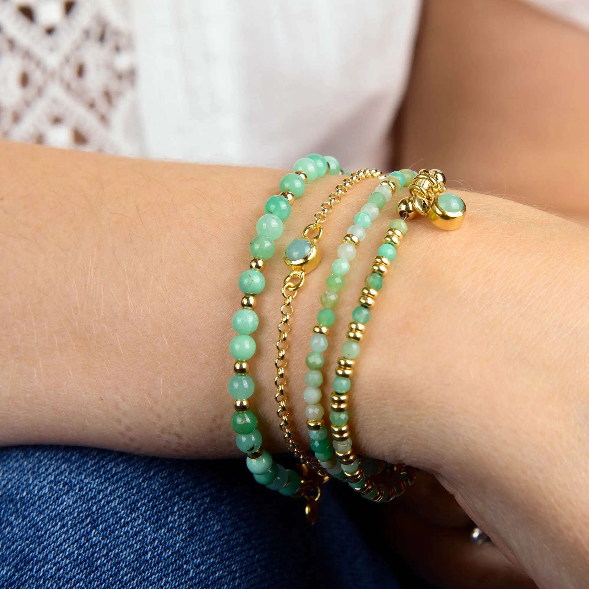 A close-up of a wrist adorned with three beaded bracelets in lush green and gold hues, including the Chrysoprase Birthstone Gem Dot Bracelet – May and a gold-plated bracelet accented with a delicate gold charm. The wearer pairs blue jeans with a white lace top, creating an elegant style that’s perfect as a May birthday gift.