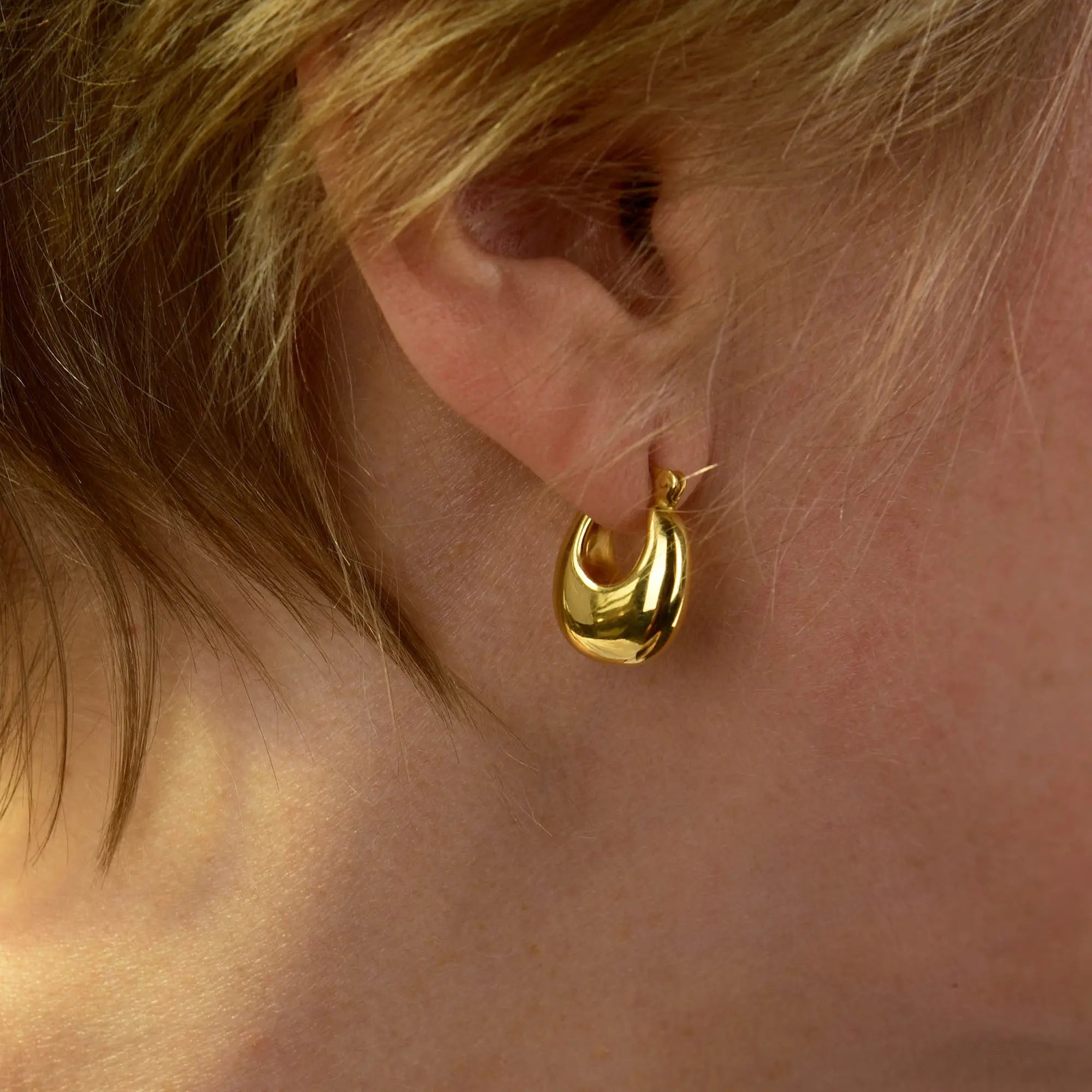 A close-up of a person’s ear adorned with the Dome Hoop Earrings. The shiny, thick gold hoops peek out from beneath light brown hair, showcasing how these sculptural earrings elevate everyday style with a touch of elegance.