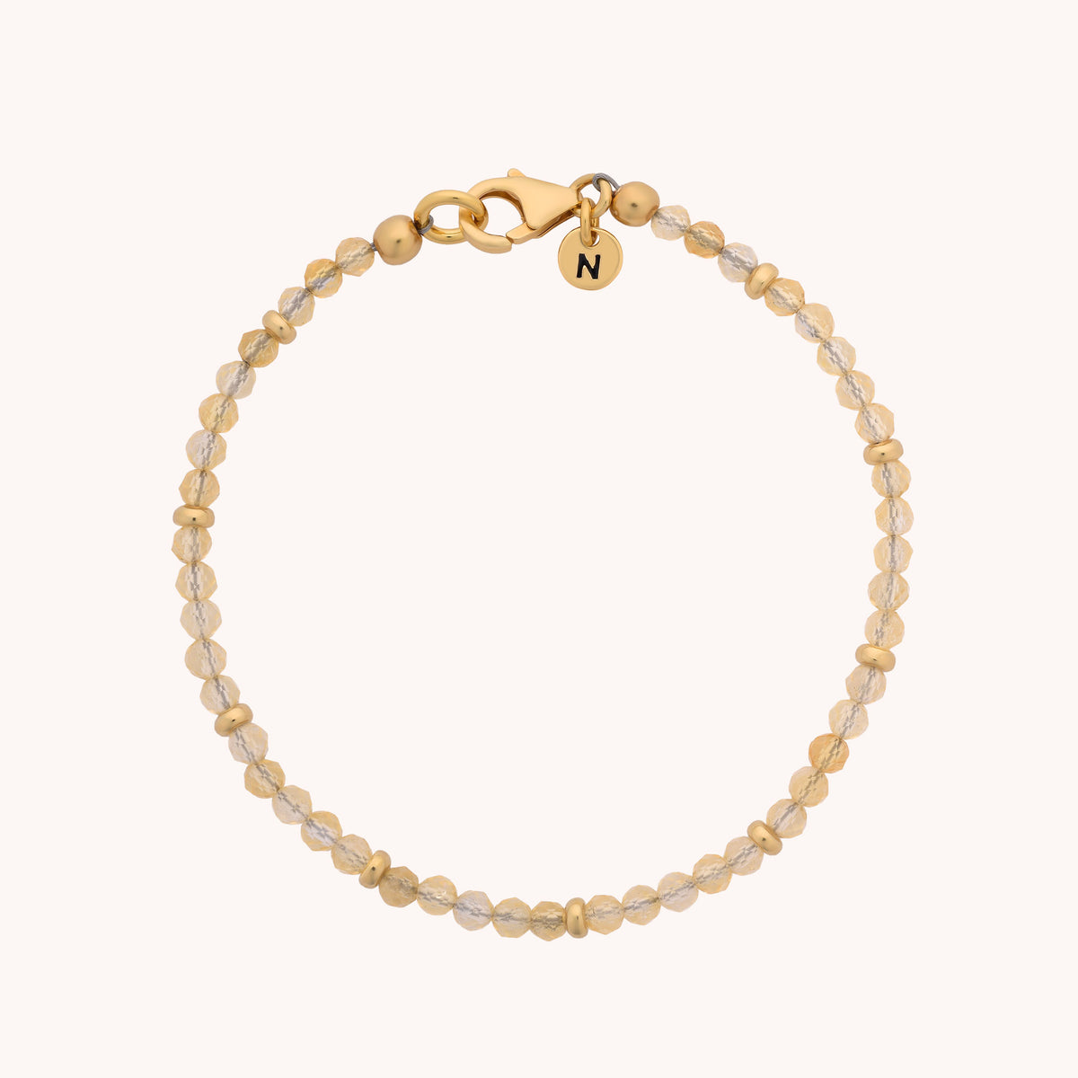Introducing the Citrine Orbit Bracelet - November: a delicate, minimalist accessory crafted from gold-plated sterling silver. This gold and clear bead bracelet features a lobster clasp and a small, round gold charm engraved with the letter N. Beautifully arranged in a circular design on a plain background, it’s the perfect birthstone piece for November.