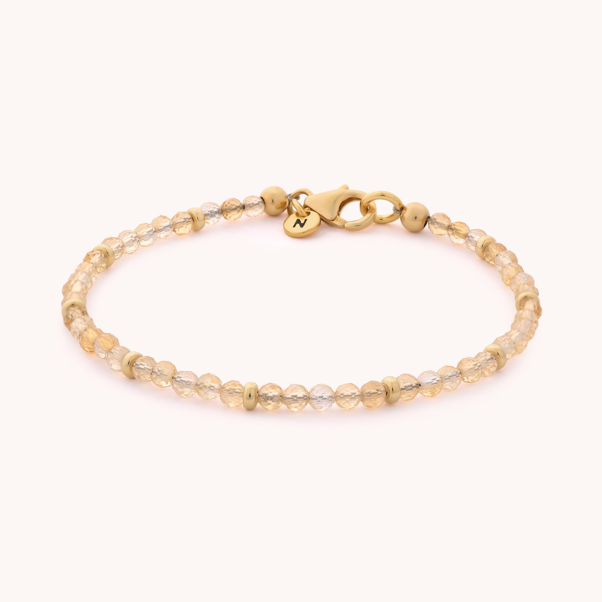 The Citrine Orbit Bracelet – November features small, faceted light brown citrine beads with gold accents, finished with a gold-plated sterling silver lobster clasp and a round charm engraved with an initial.