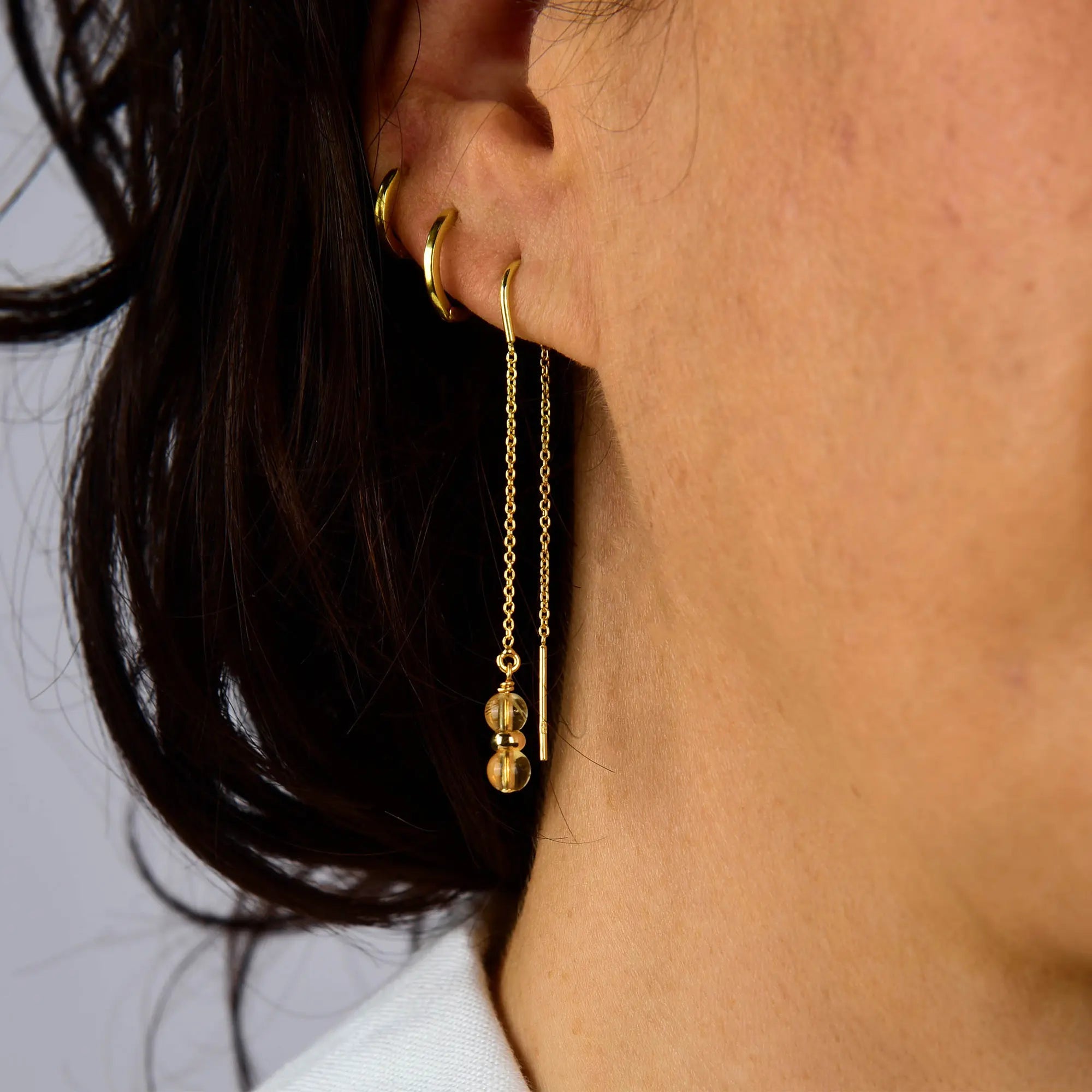 Close-up of a person’s ear adorned with three small gold hoop earrings and the Citrine Fall Earrings Birthstone November—a long gold threader earring featuring two spherical gold beads—against a neutral background.
