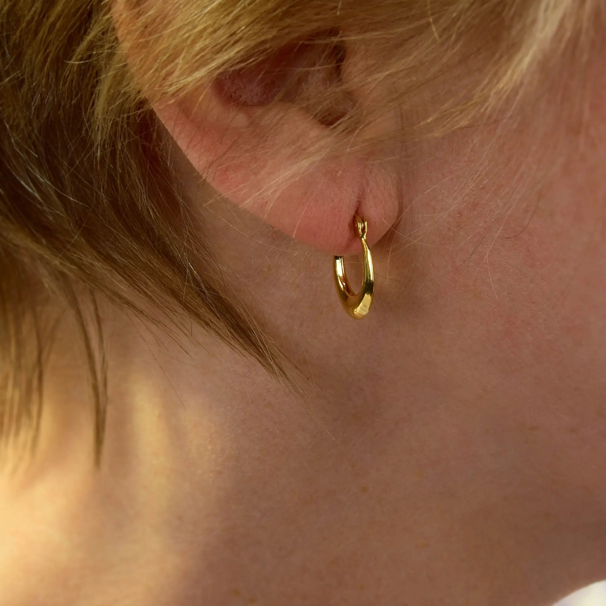Close-up of a person’s ear adorned with a small, shiny gold Contour Hoop Earrings. Light brown hair partially covers the ear, and the background is softly blurred.
