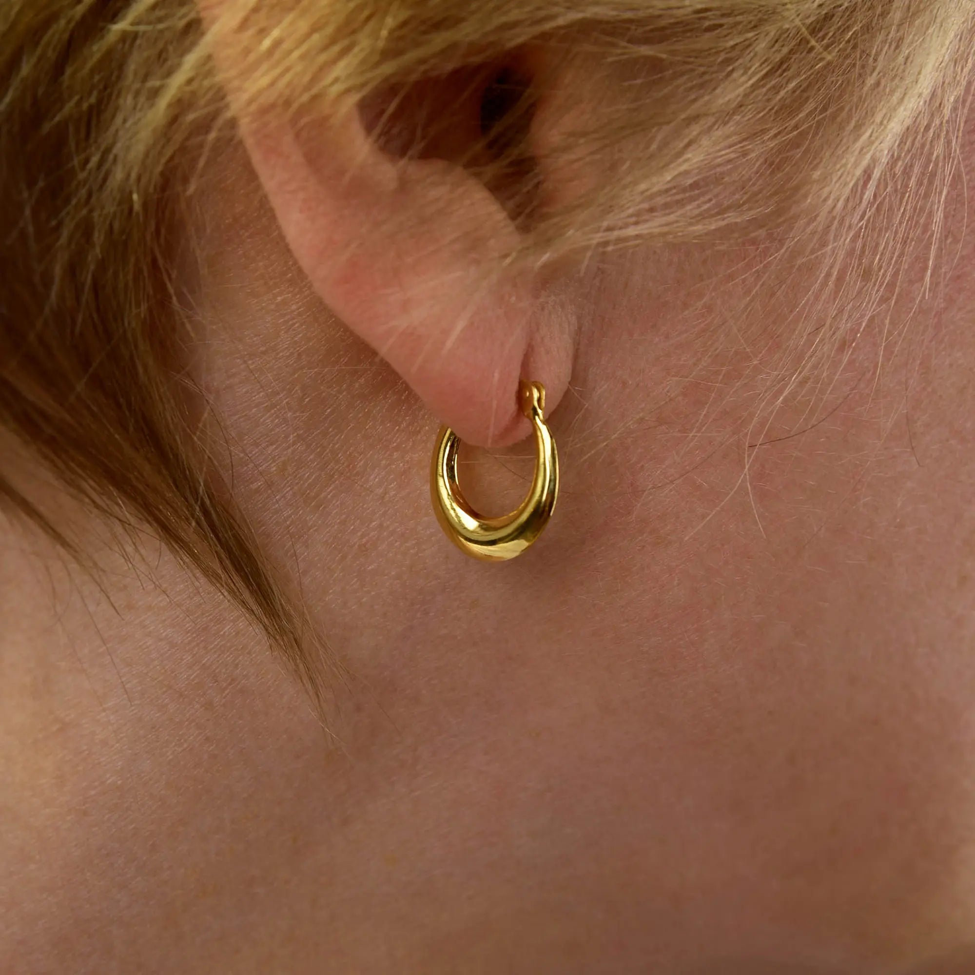 Close-up of a person’s ear adorned with the Contour Hoop Earring, a small and shiny gold piece that echoes a sculptural design. The person’s light brown hair is partially draped over the top of the ear.