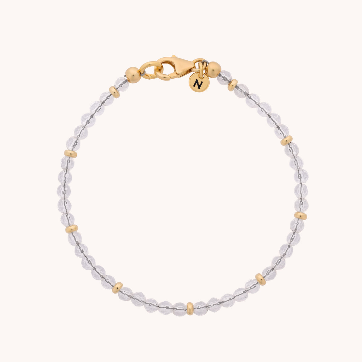 Introducing the Rock Crystal Orbit Bracelet - April: a delicate accessory featuring clear round rock crystal beads, small gold spacers, a gold clasp, and a petite gold charm engraved with the letter N.
