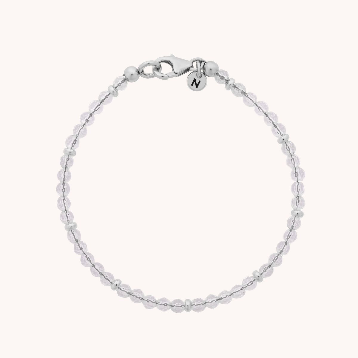 Introducing the Rock Crystal Orbit Bracelet - April: a delicate sterling silver bracelet adorned with small round beads, a secure lobster clasp, and a tiny circular charm engraved with the letter N. Beautifully displayed on a light background, this elegant piece makes a thoughtful gift or charming accessory for any occasion.