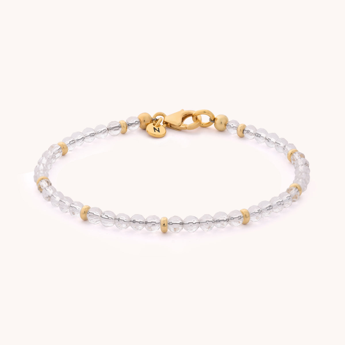 Rock Crystal Orbit Bracelet - April: Features clear rock crystal beads accented with small gold spacer beads, a gold clasp, and a small round gold charm engraved with the letter N, photographed on a plain white background—a subtle nod to the April birthstone.