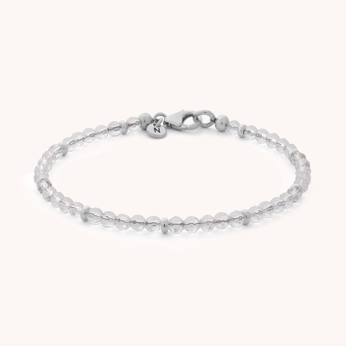 The Rock Crystal Orbit Bracelet - April features small, round, clear rock crystal beads and a sterling silver clasp with a petite round tag near the closure, elegantly displayed on a light background.