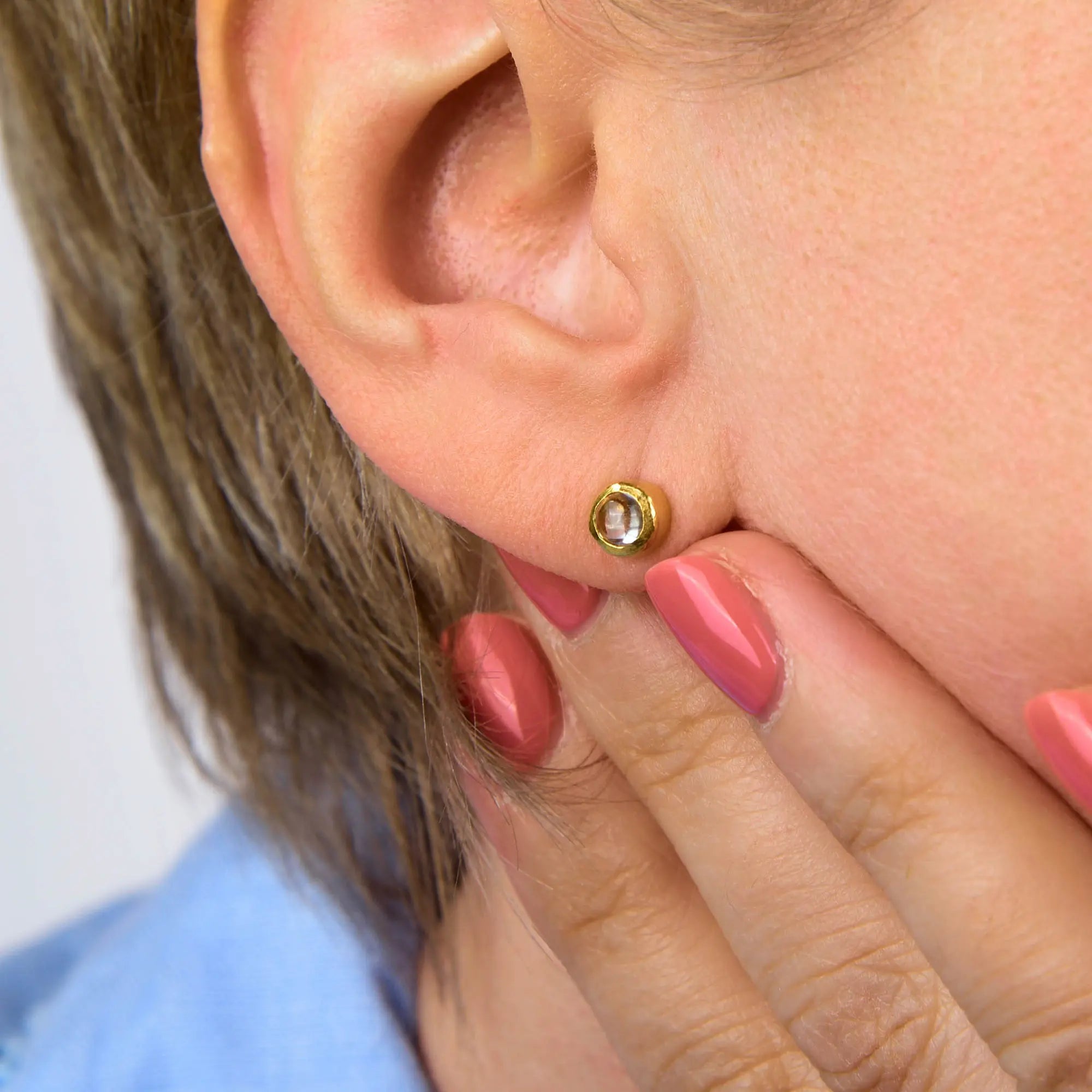 A close-up of a person’s ear adorned with the Crystal Mini Gem Dot Birthstone Earrings for April in sterling silver. The person has short hair and is touching their ear, showing off pink-painted nails.