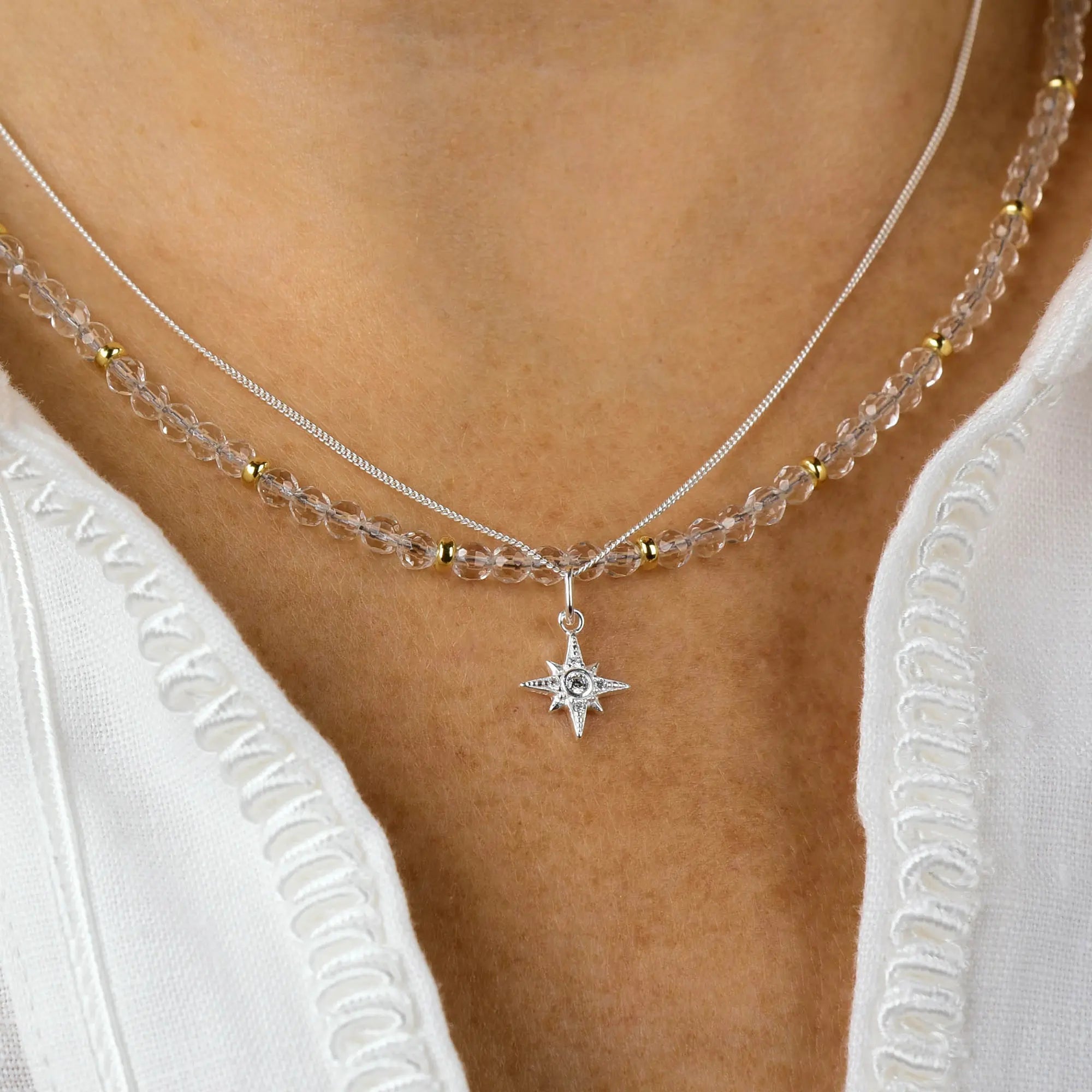 A close-up of a person wearing a layered necklace: one features clear beads and gold accents, while the other is the Celestial Star Necklace—a silver chain with a star-shaped pendant—styled over a white textured blouse.