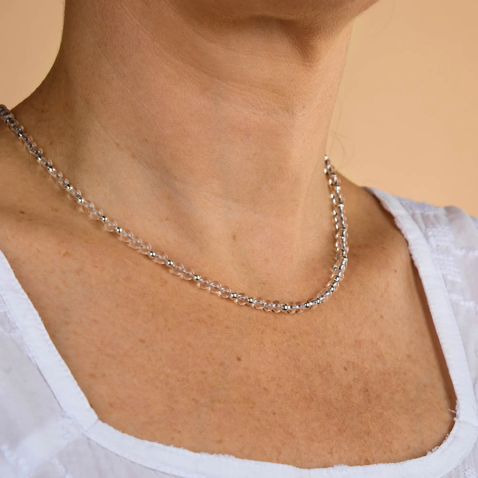 A person wearing a white top and the Clear Quartz Jewel Bead Birthstone Necklace - April, shown from the shoulders up against a beige background.