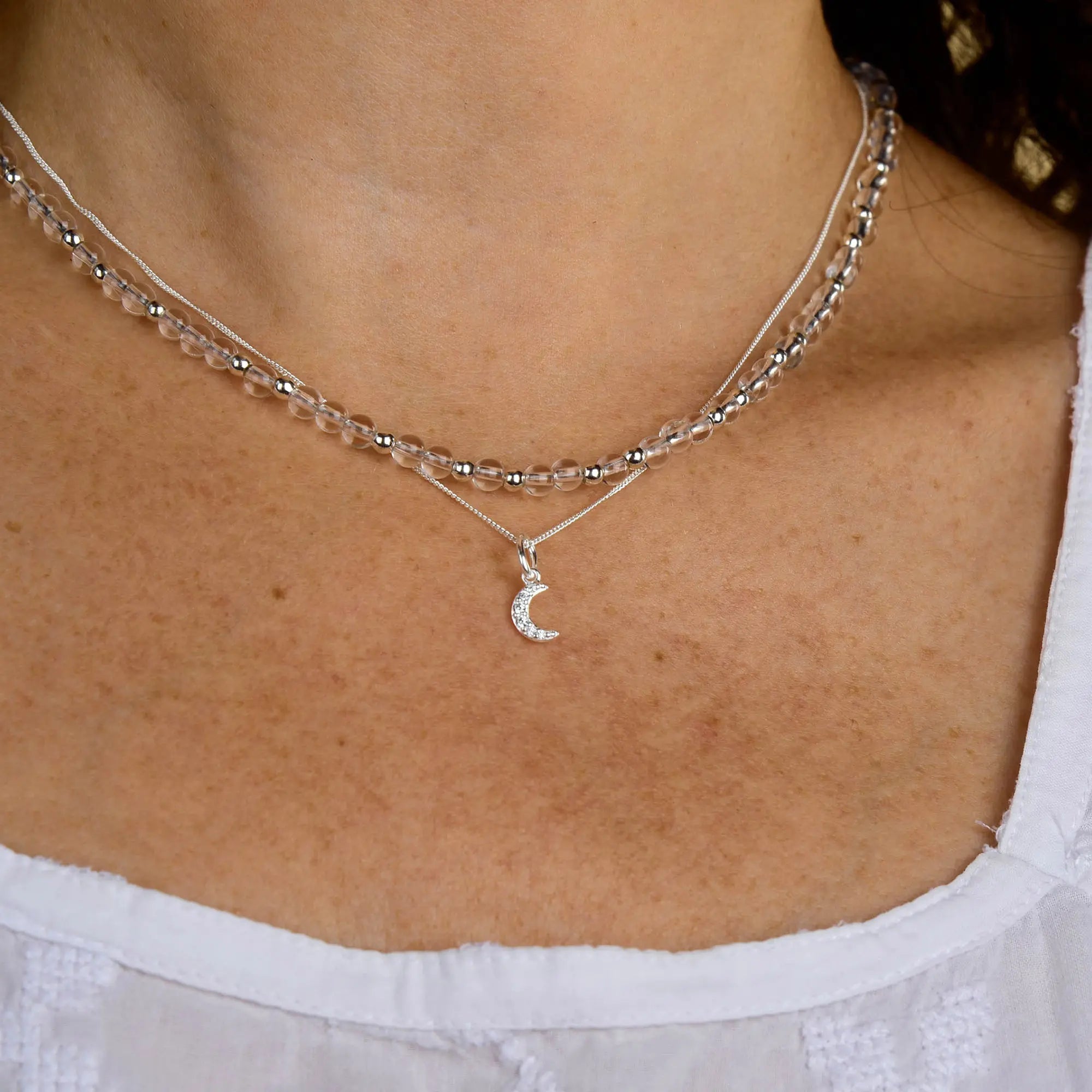 A close-up of a person wearing two silver necklaces: one features the Clear Quartz Jewel Bead Birthstone Necklace - April with natural gemstone beads, and the other is adorned with a crescent moon pendant. The person is dressed in a white top.