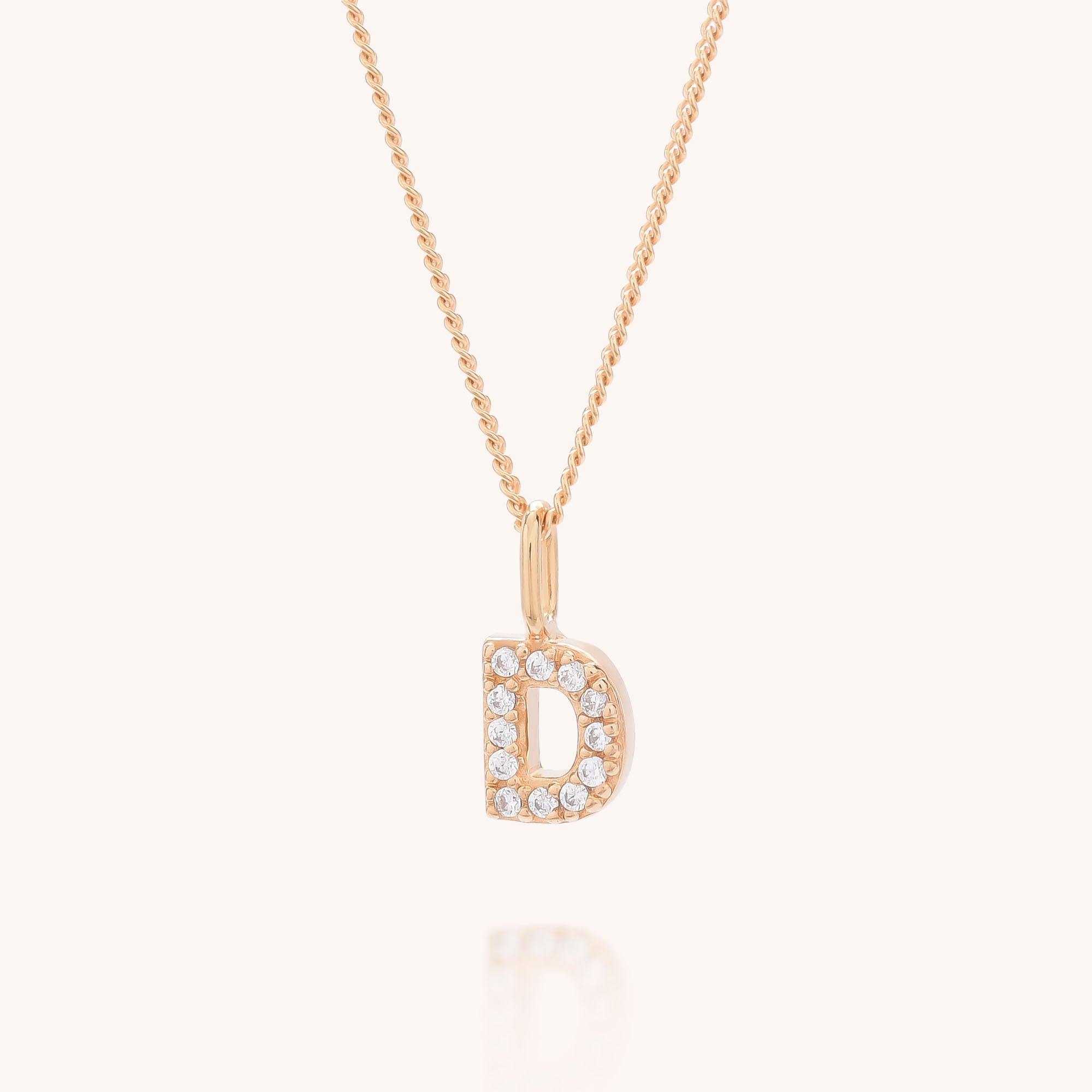 The Initial Sparkle Necklace features a gold pendant shaped like the letter D, adorned with sparkling cubic zirconia, and is showcased on a light beige background.