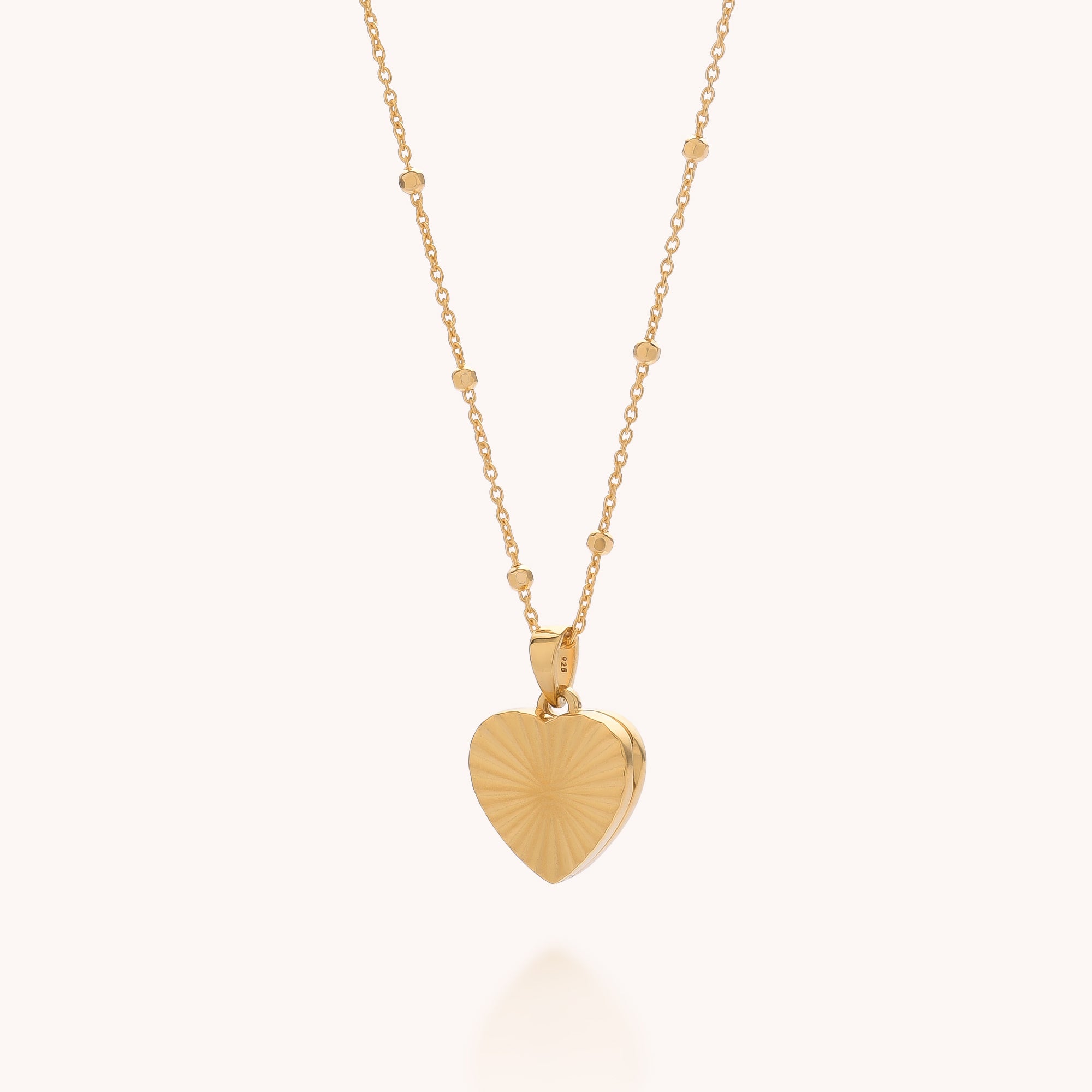 The Aura Heart Locket Necklace features a heart-shaped locket with a subtle sunburst engraving on a light background and gold-plated small bead accents along the chain.