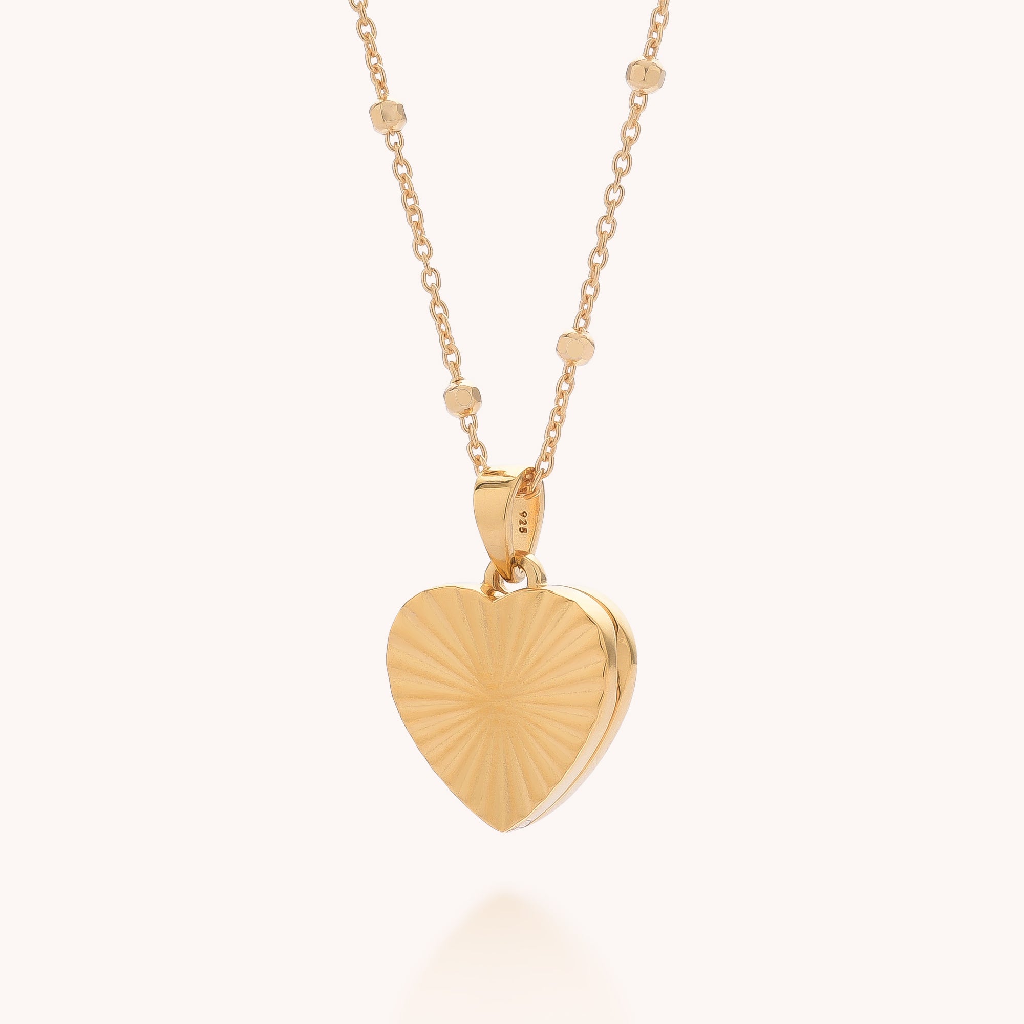 The Aura Heart Locket Necklace features a delicate gold plated chain with small beads and a heart-shaped locket engraved with a sunburst pattern, displayed on a light background.