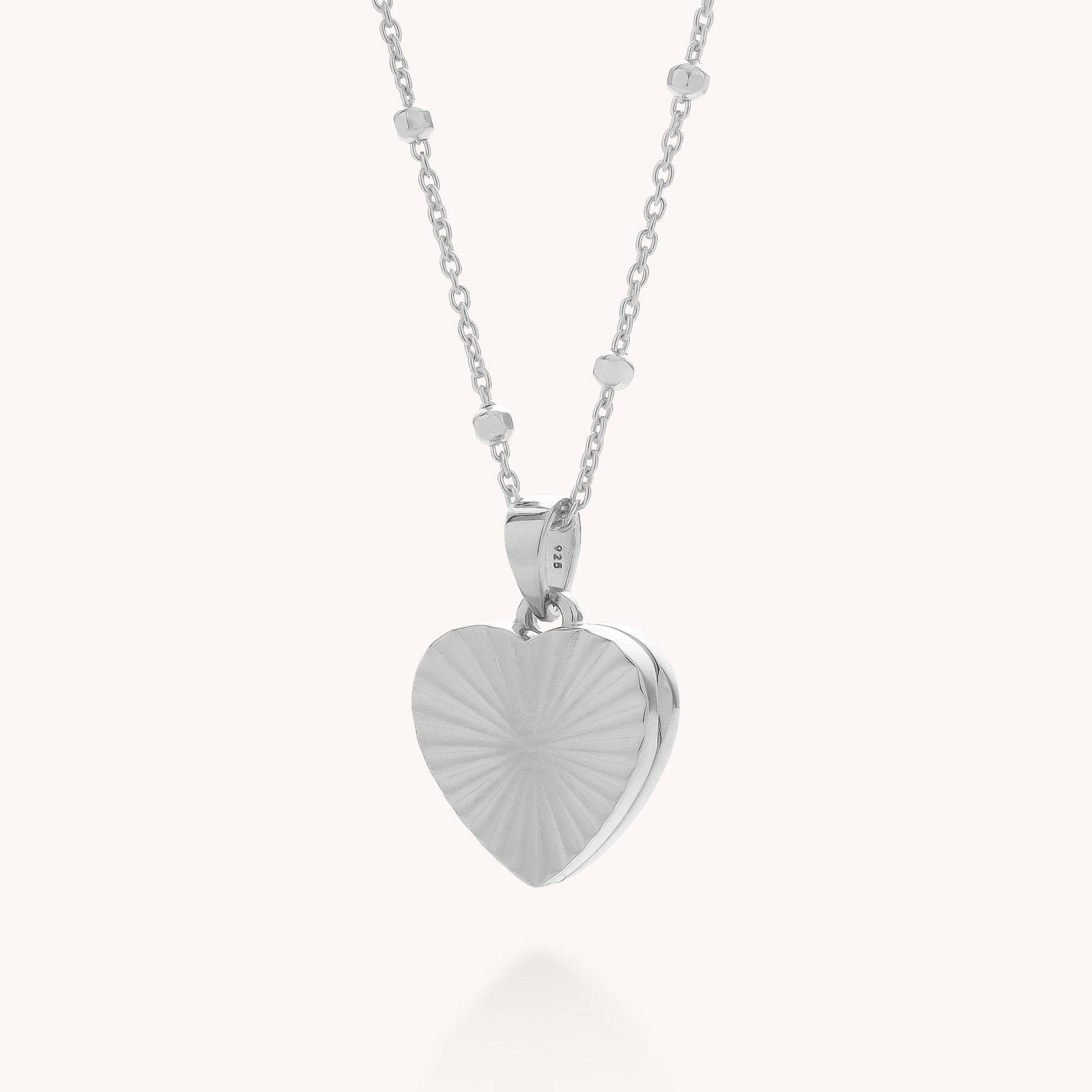 The Aura Heart Locket Necklace features a delicate silver chain with small beads and a heart-shaped pendant engraved with a sunburst pattern—an elegant, timeless accessory.