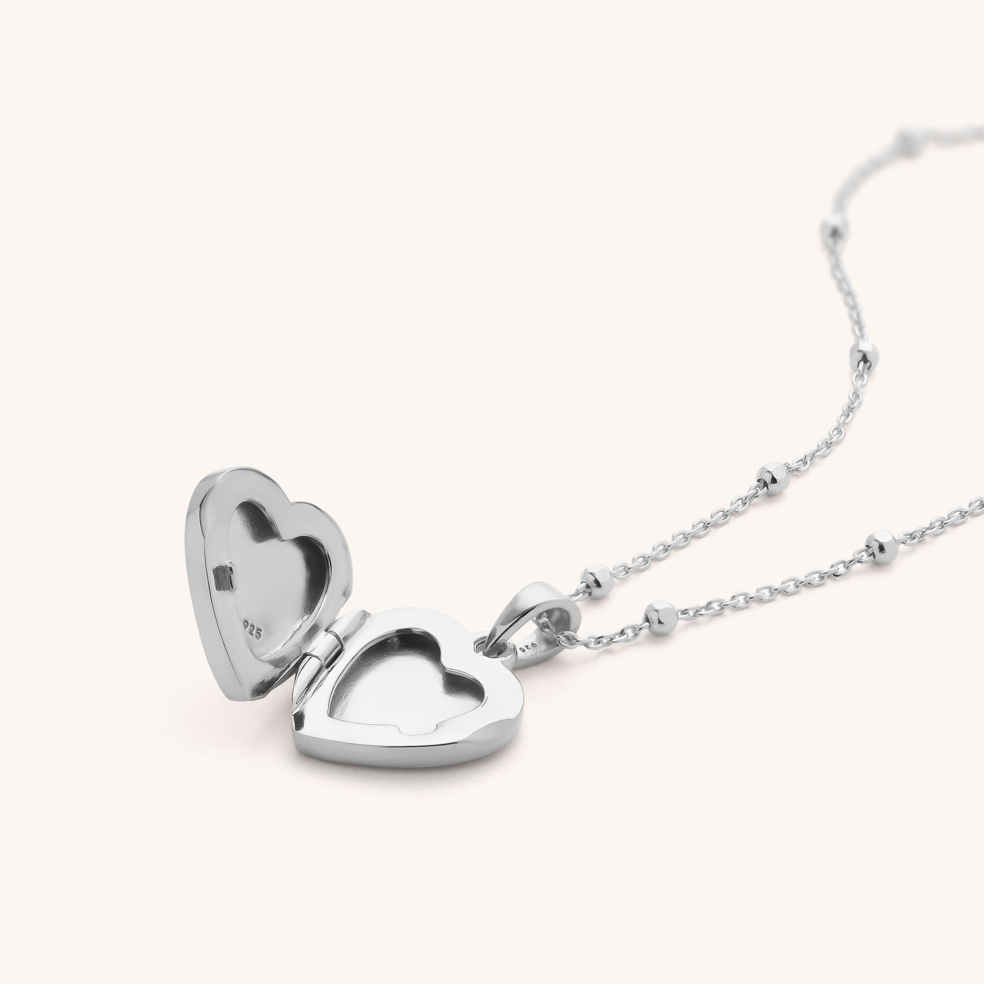 The Aura Heart Locket Necklace features a sterling silver heart-shaped pendant on a delicate chain, shown open to reveal two empty compartments inside, set against a plain light background.