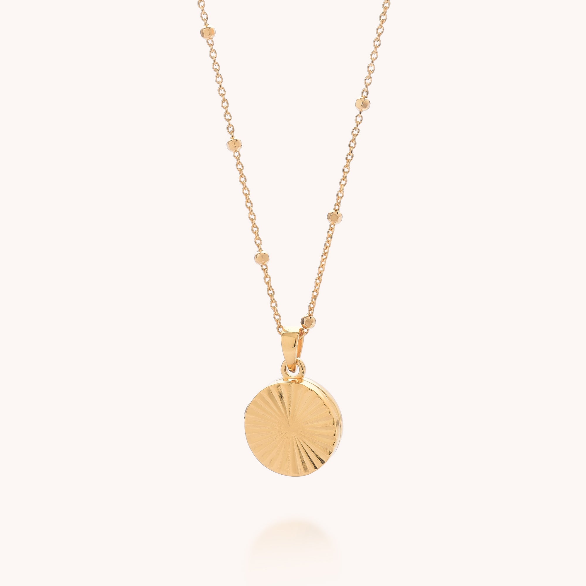 The Aura Round Locket Necklace features a round, textured pendant with a subtle radiating pattern on a beaded chain, shown against a light background.