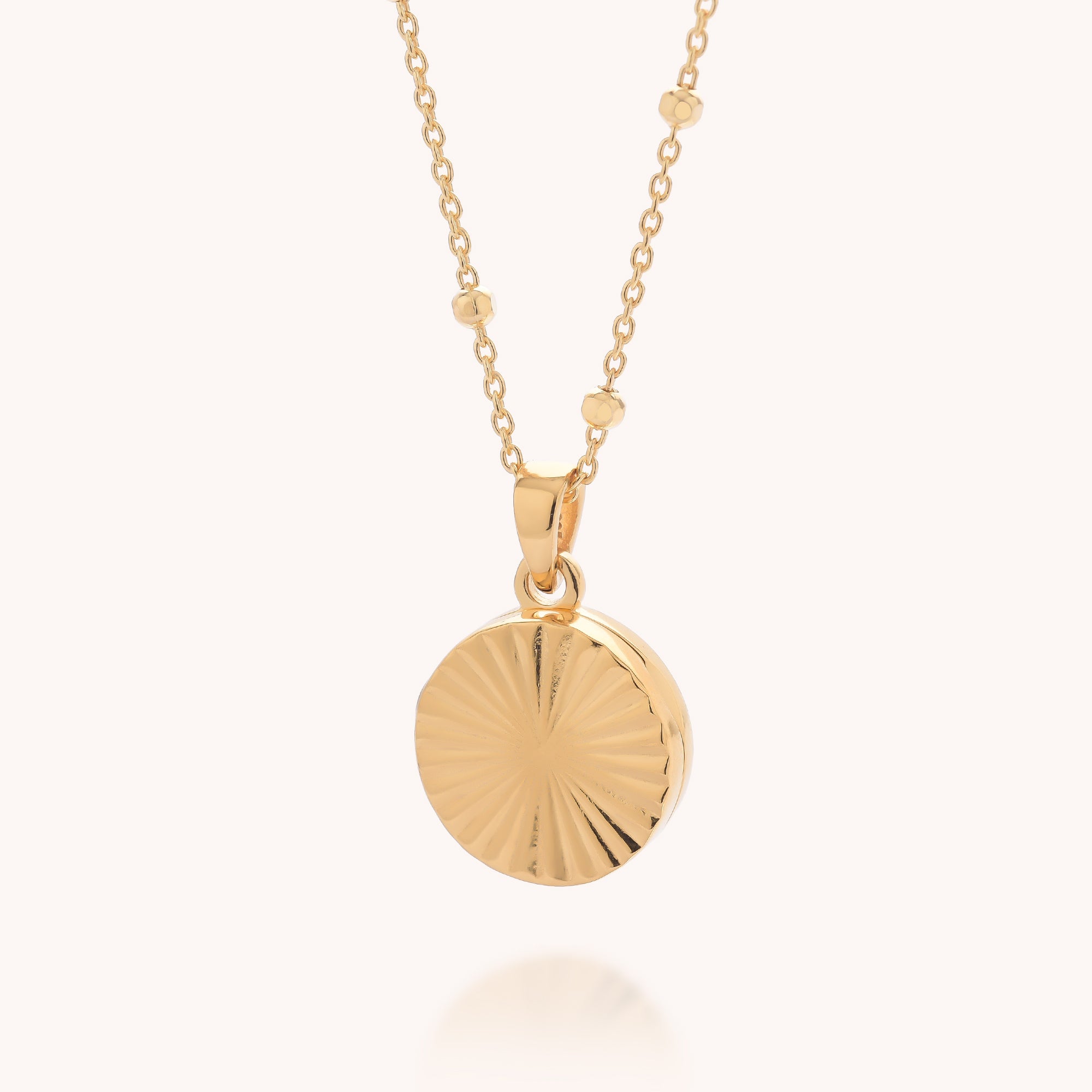 The Aura Round Locket Necklace features a gold chain with small beads and a round, textured sunburst pendant for an elegant touch.
