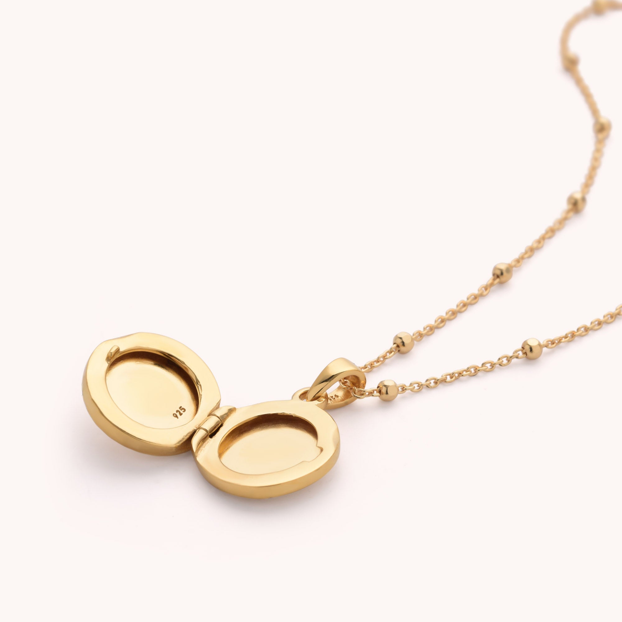 The Aura Round Locket Necklace is shown open, revealing two empty oval compartments. It features a delicate chain with small, evenly spaced beads.