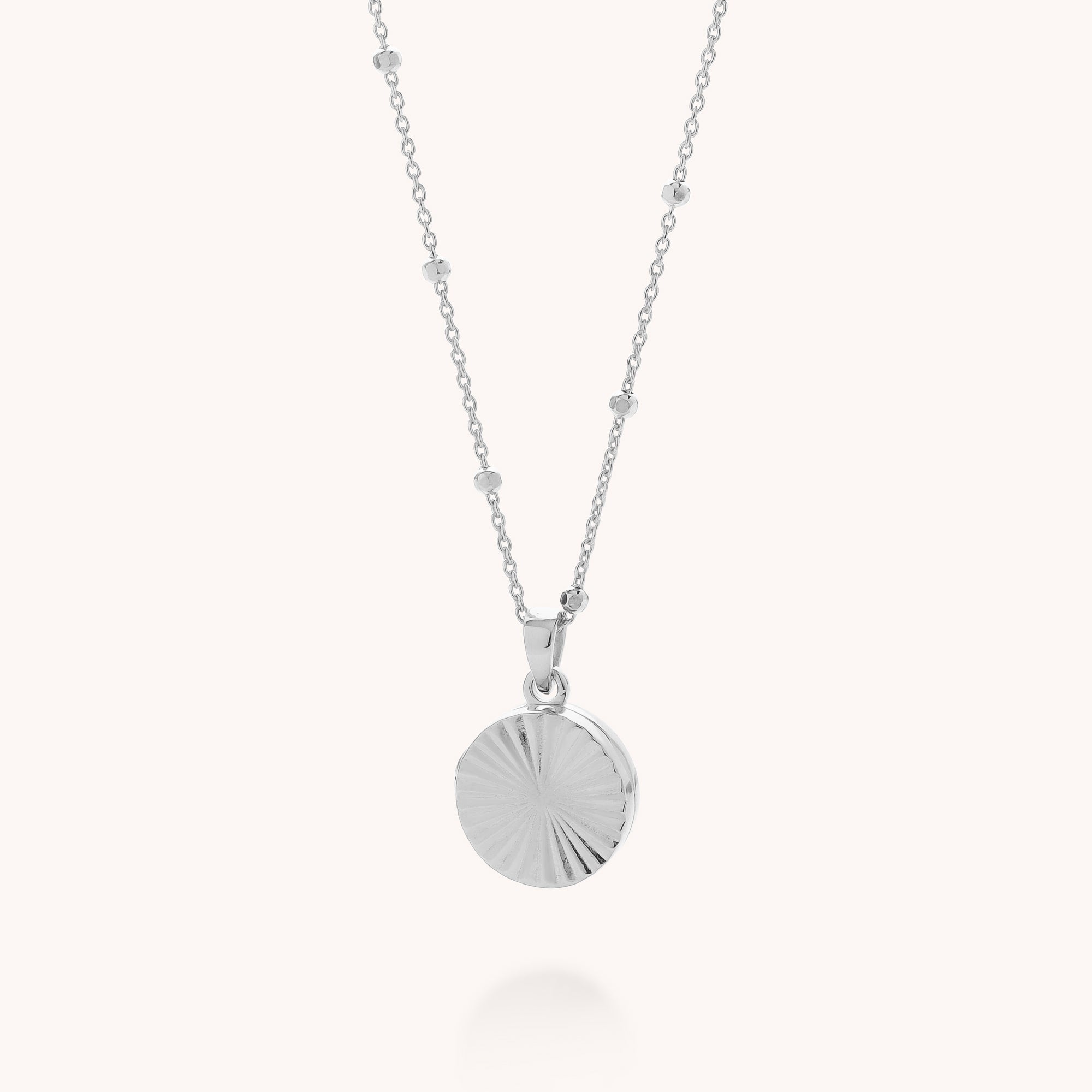 The Aura Round Locket Necklace is a delicate sterling silver piece with small bead accents and a round, textured sunburst pendant, shown against a light background.