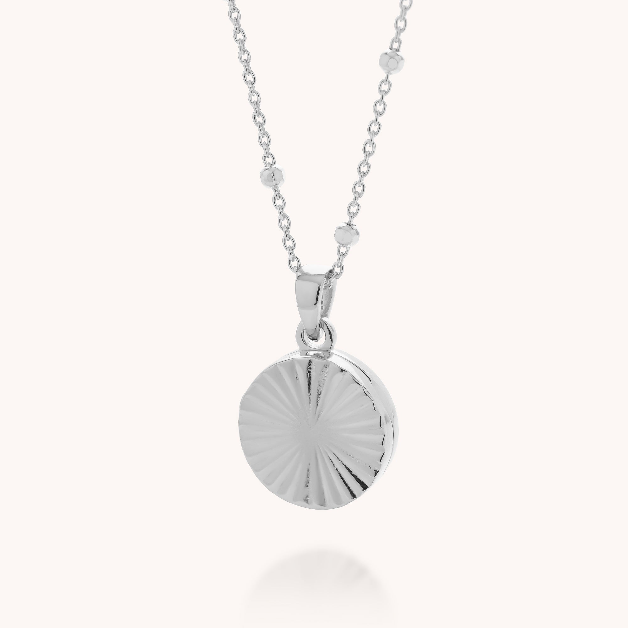 The Aura Round Locket Necklace features a round, textured pendant with a sunburst pattern in sterling silver, hanging from a delicate chain accented with small decorative beads.