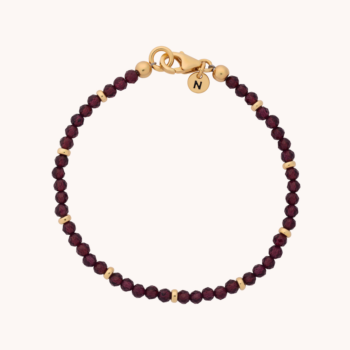 Introducing the Garnet Orbit Bracelet - January: Featuring a gold clasp, small round deep red garnet beads reminiscent of the January birthstone, and gold accent beads. A round gold charm engraved with the letter N adds a personal touch. This elegant bracelet is perfect for celebrating special moments.