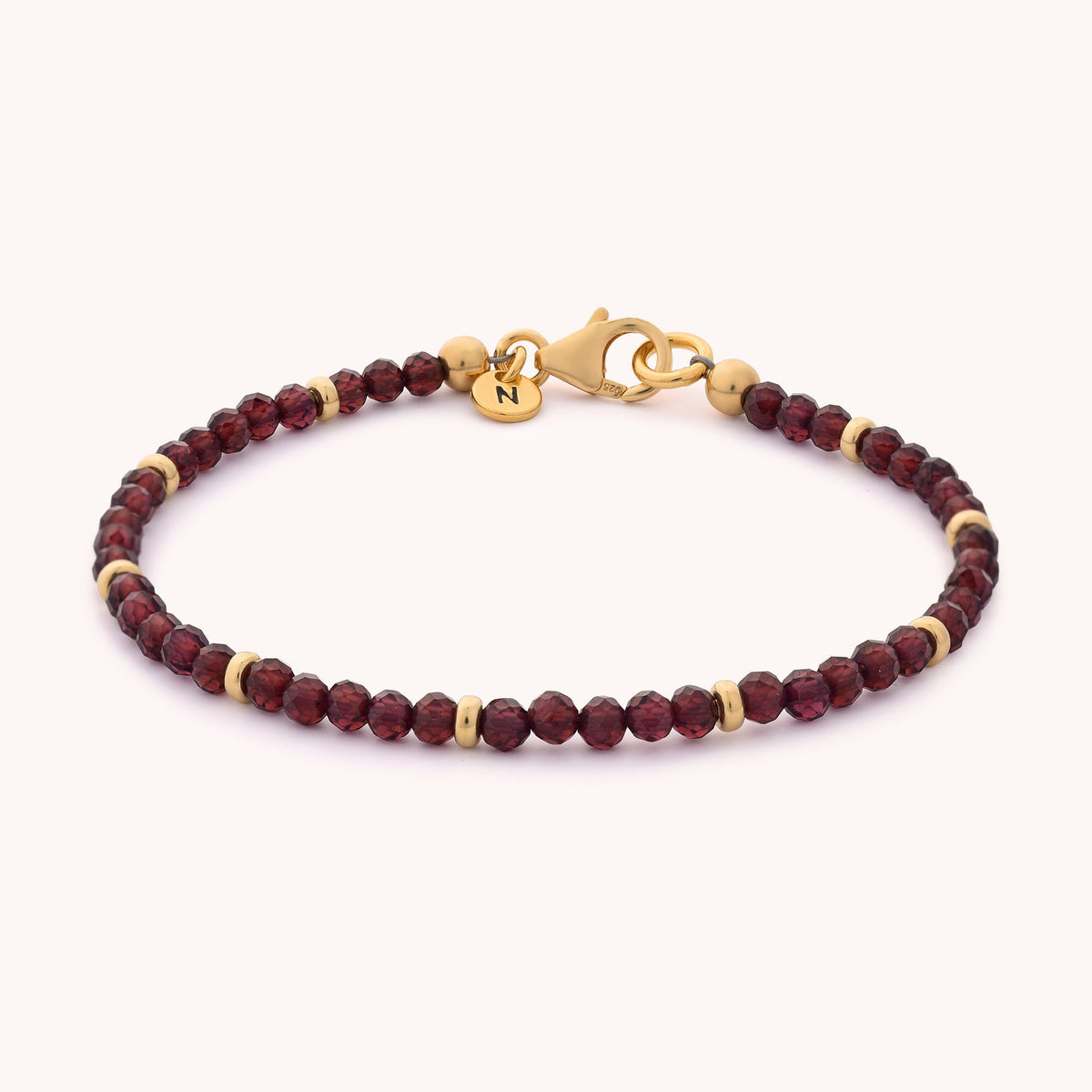 Introducing the Garnet Orbit Bracelet - January: a delicate piece adorned with small, round deep red garnet beads and elegant gold accents. It features a gold lobster clasp and a petite gold charm engraved with the letter N, making it the perfect accessory to celebrate a January birthstone in style.