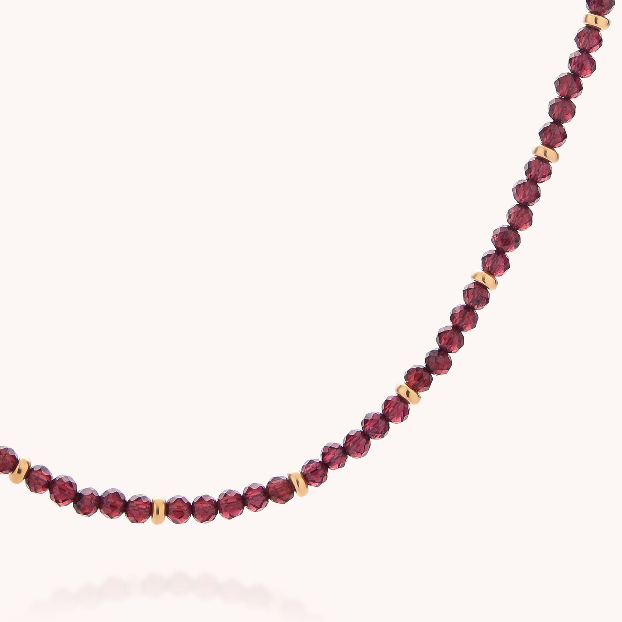 A close-up of the Garnet Orbit Necklace showcases small, faceted dark red garnet beads interspersed with gold spacer beads, beautifully displayed on a plain light background.