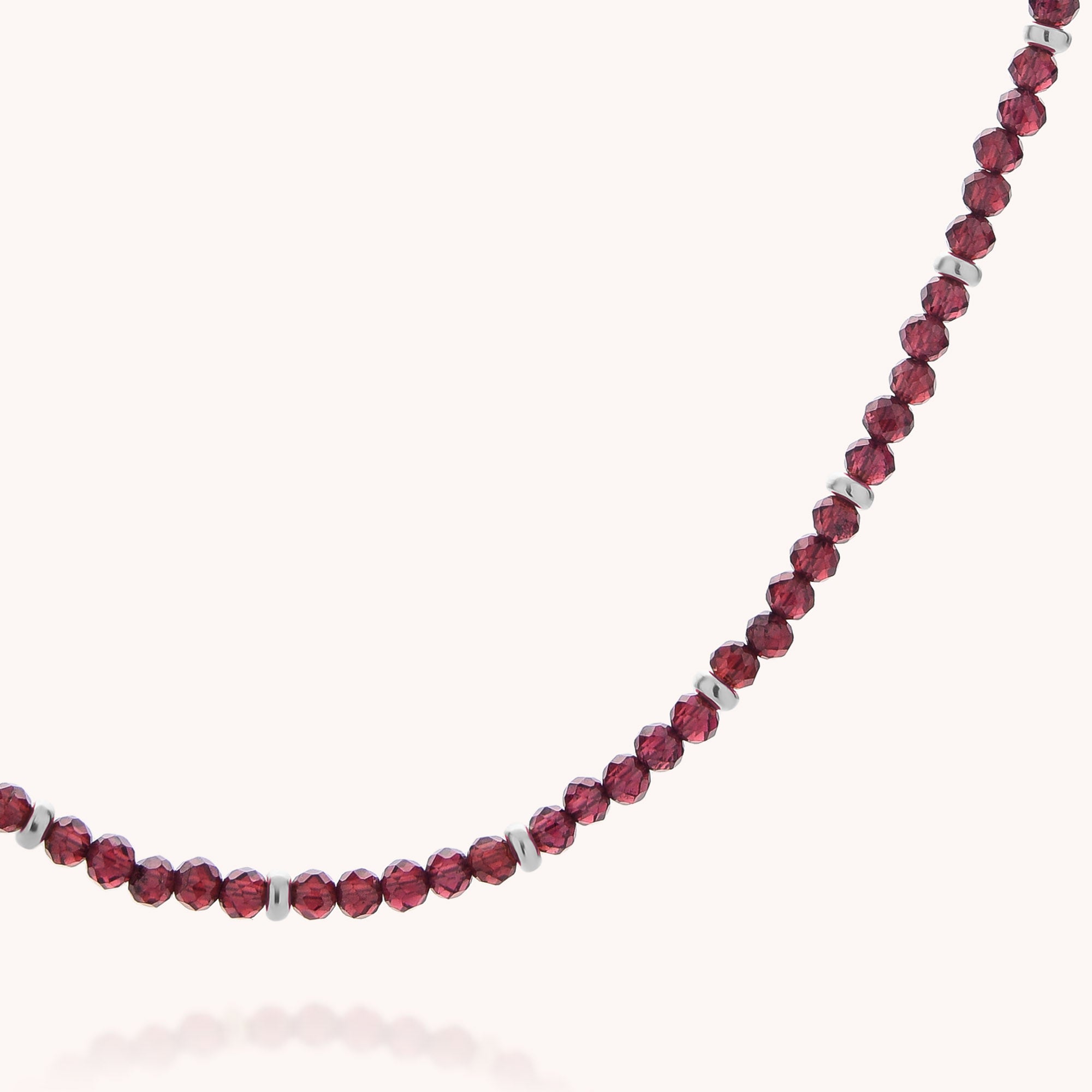 A close-up of the Garnet Orbit Necklace showcases small, faceted, deep red garnet beads accented with delicate silver spacers, elegantly arranged in a curved line on a plain light background—a sophisticated piece of handcrafted jewelry.