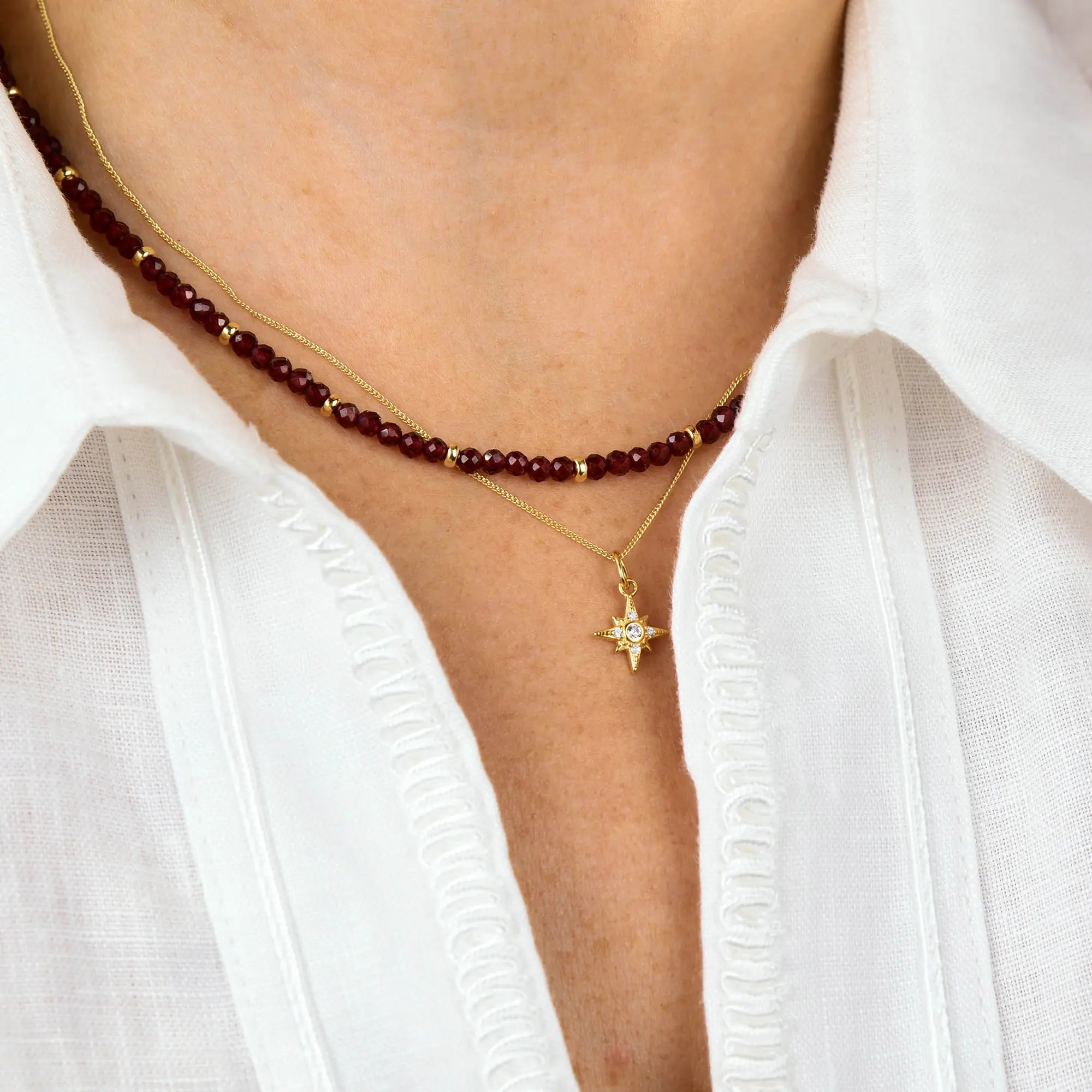 A person wearing a white collared shirt with two necklaces: one is a gold chain with a star-shaped pendant, while the other is the Garnet Orbit Necklace, adding an elegant touch of artisanal jewelry to the look.