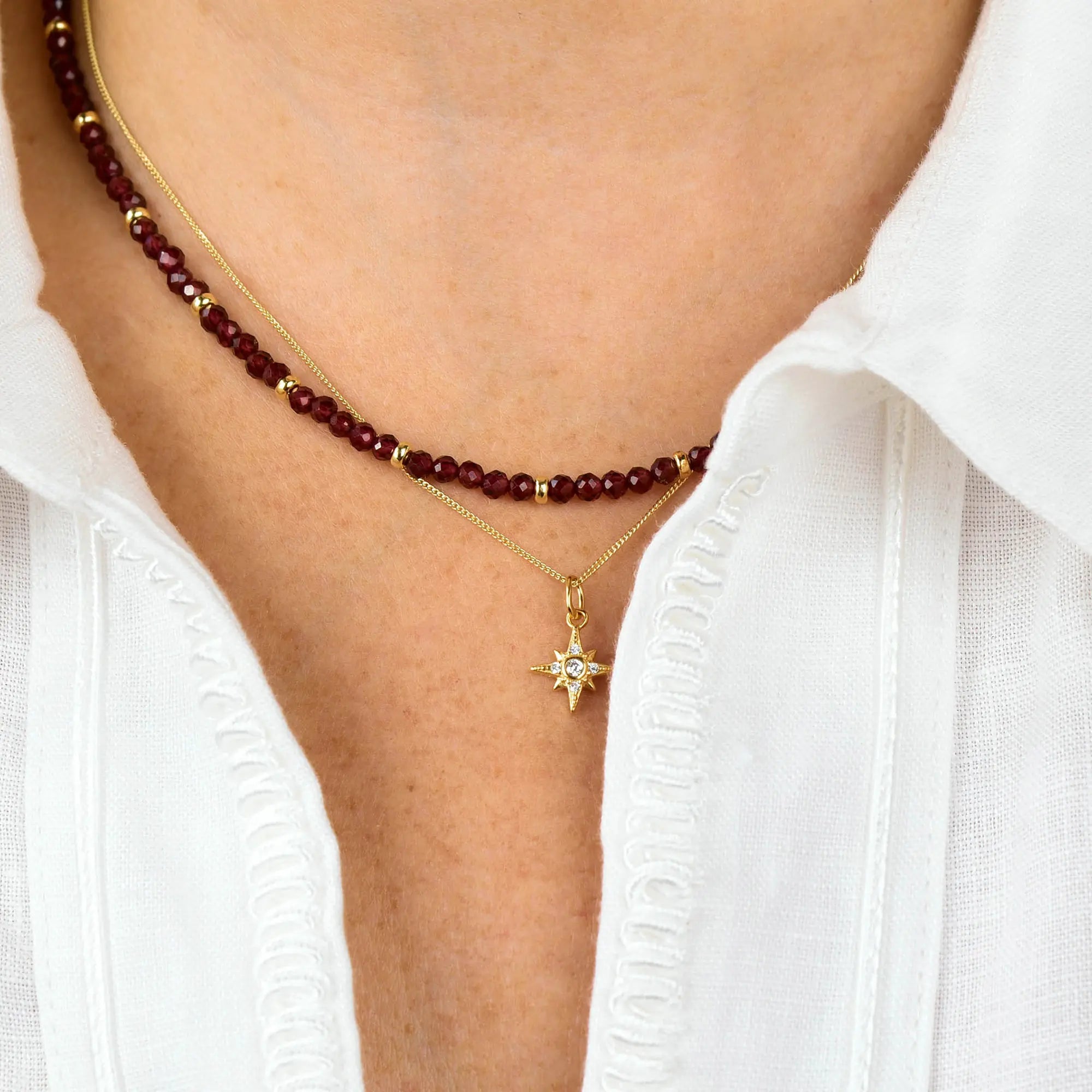 A person wearing a white shirt and two necklaces: one is the Garnet Orbit Necklace, featuring handcrafted red garnet beads, and the other is a gold chain birthstone necklace with a star-shaped pendant adorned with a small gem in the center.