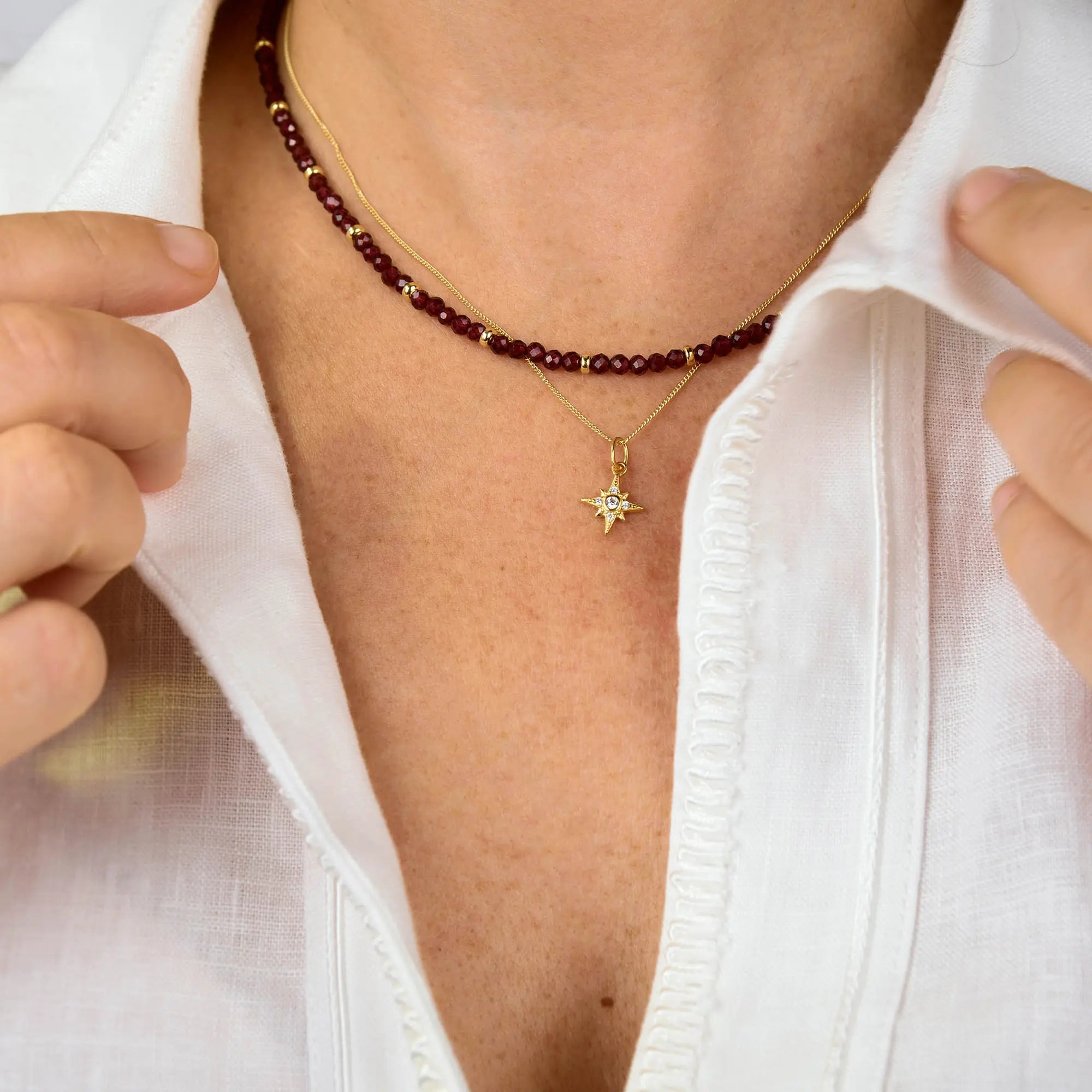 A person wearing a white shirt displays two necklaces: one is the Garnet Orbit Necklace, featuring small dark red beads for a handcrafted jewelry look reminiscent of delicate garnet stones; the other is a gold chain with a star-shaped pendant adorned with a small gemstone, evoking the charm of a birthstone necklace.