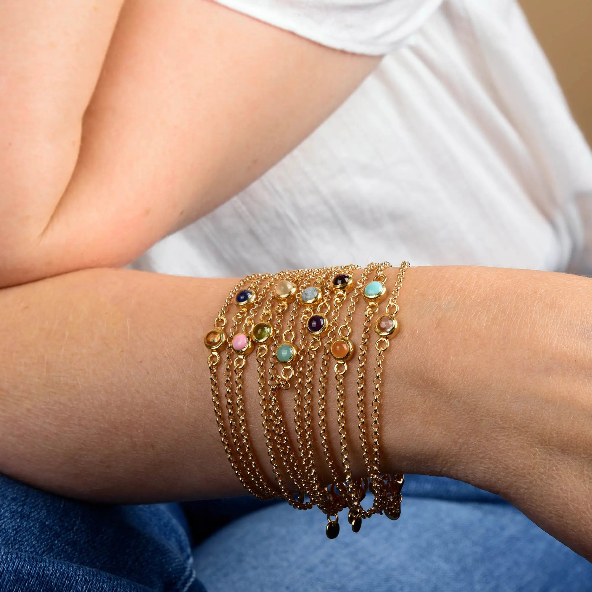 A person wearing multiple gold chain bracelets, including the Peridot Birthstone Gem Dot Bracelet - August adorned with colorful gemstone charms, rests their arm on their knee. The person is dressed in a white top and blue jeans.