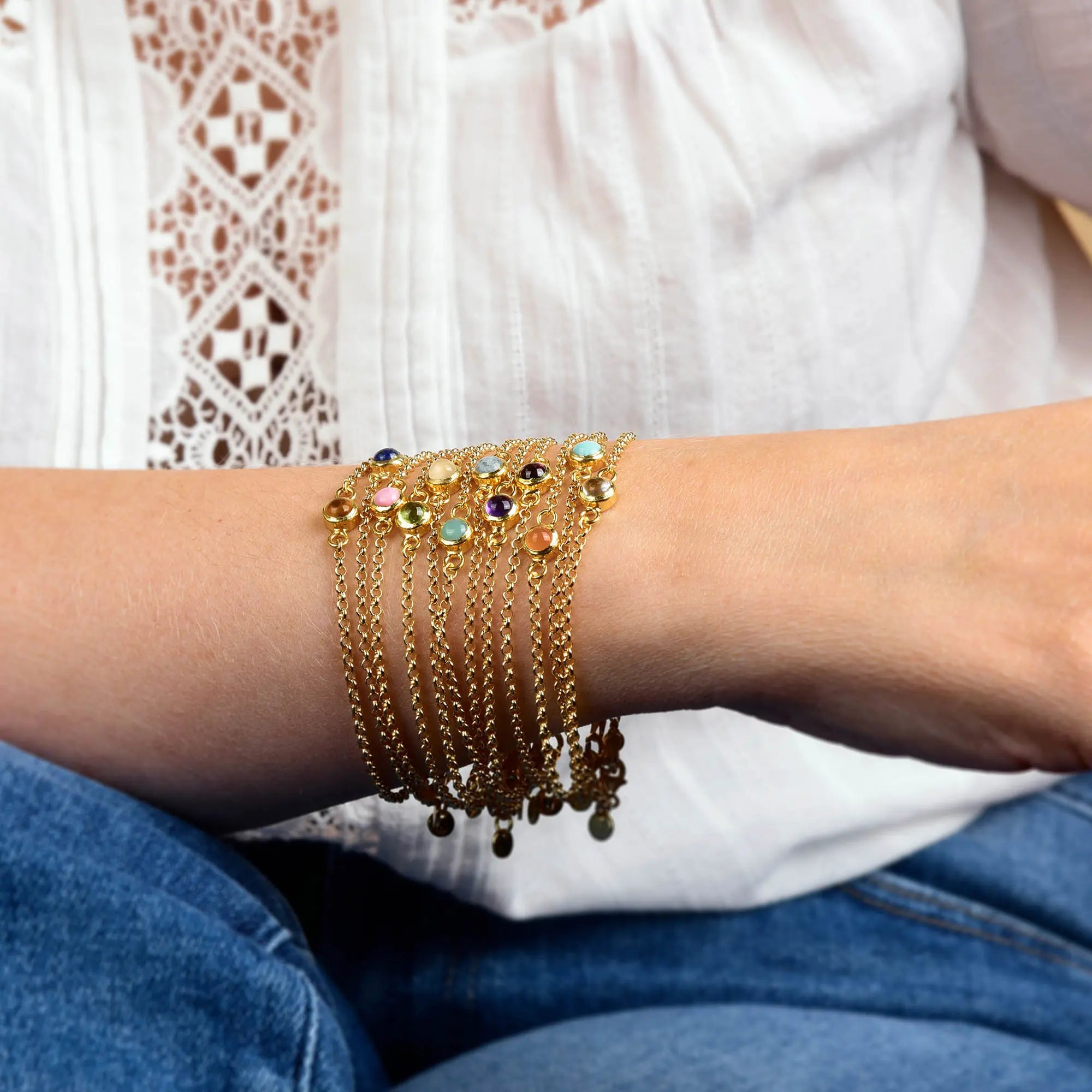 A person wearing several gold chain bracelets, including the Aquamarine Birthstone Gem Dot Bracelet – March, which makes a thoughtful March birthday gift, rests their arm on their knee. They are dressed in a white lace-patterned blouse and blue jeans.