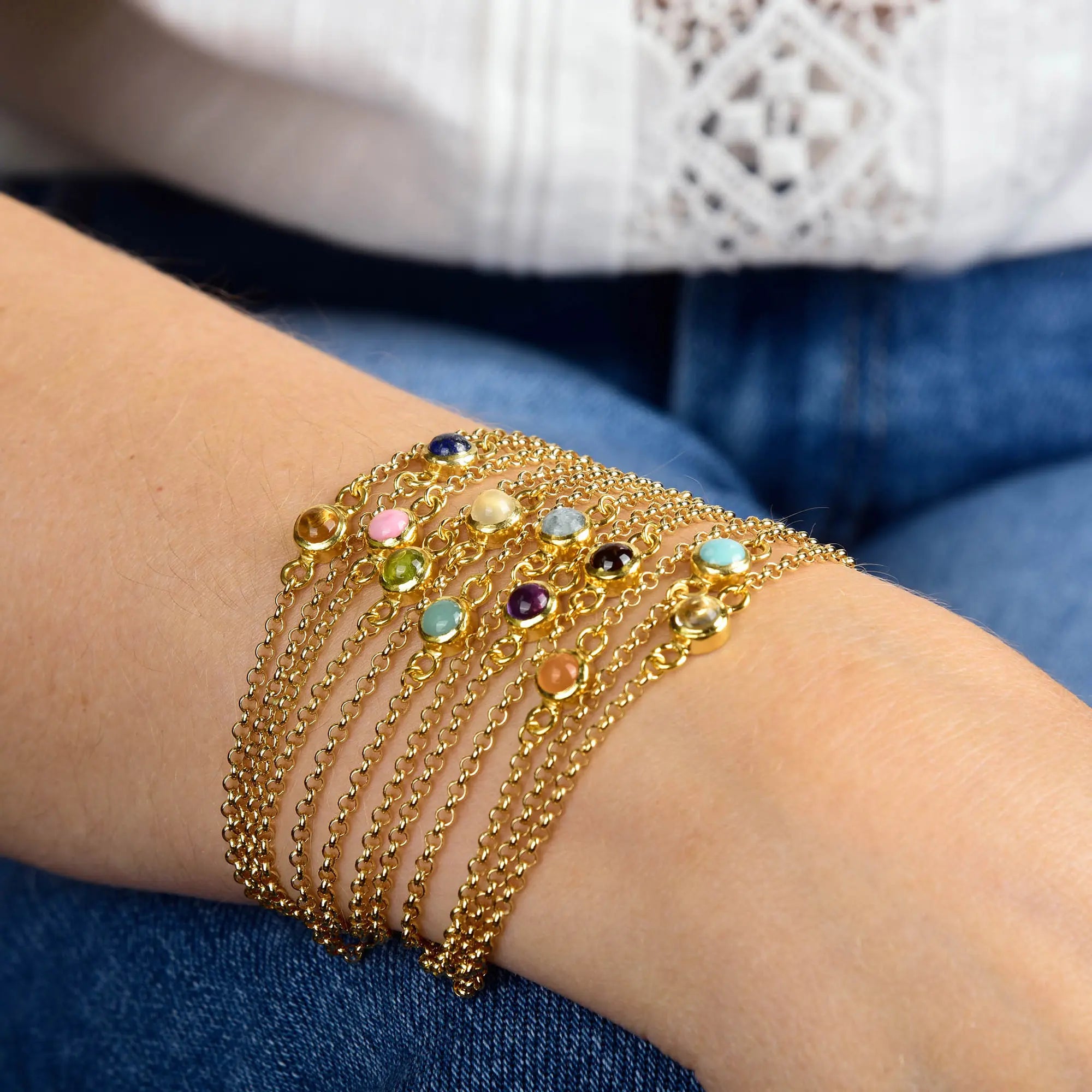 A person wearing the Carnelian Birthstone Gem Dot Bracelet - July—a multi-strand gold plated bracelet featuring vibrant gemstones, including a prominent Carnelian birthstone—rests it on their wrist while dressed in blue jeans and a white patterned top.