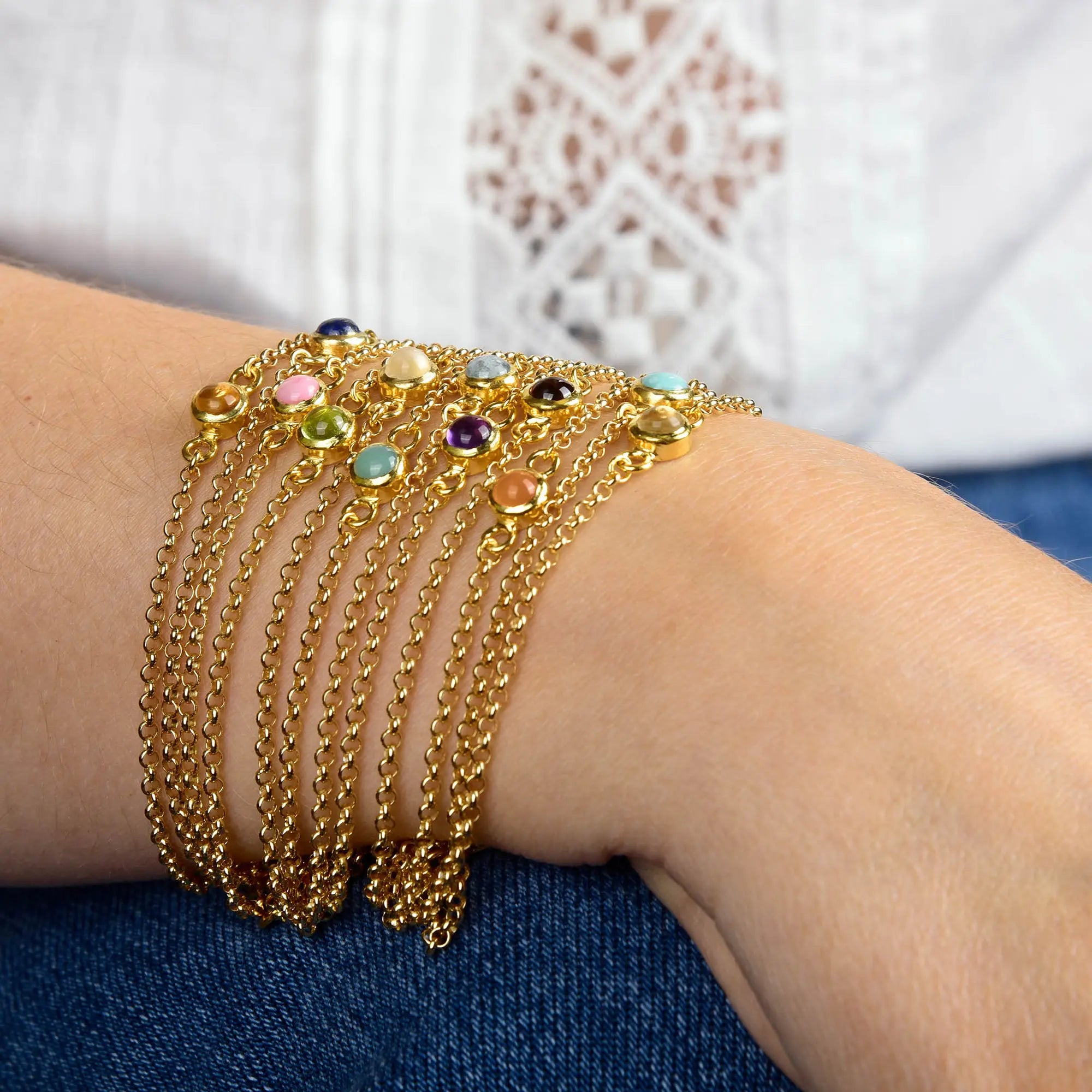 A wrist styled with multiple gold chain bracelets, including the elegant Citrine Birthstone Gem Dot Bracelet - November, each adorned with vibrant round gemstones. The wearer pairs a white patterned blouse with blue denim, drawing attention to the citrine gem on their sophisticated gold plated November birthstone bracelet.