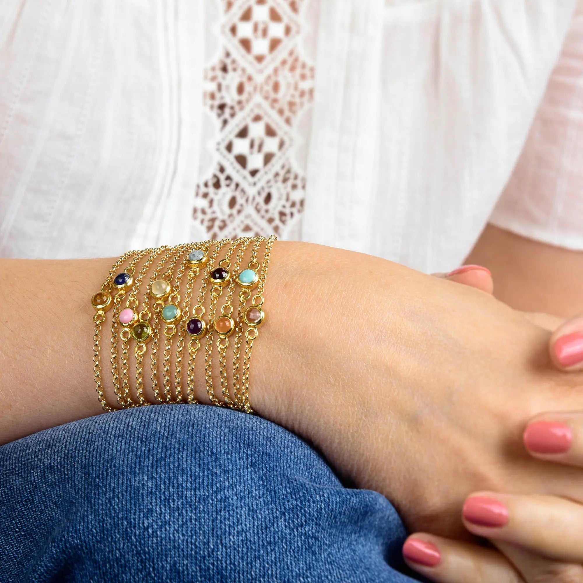A person wearing blue jeans and a white lace top rests their hand on their knee, showing multiple gold chain bracelets, including the Turquoise Birthstone Gem Dot Bracelet - December adorned with colorful gemstone charms, and pink nail polish.