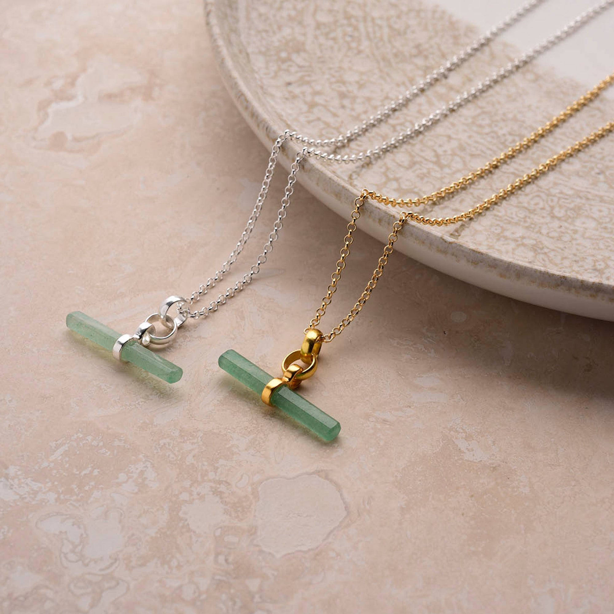 Two Gemstone T Bar Necklaces—one with a green bar on a silver chain, the other on gold—are displayed on light beige marble beside a speckled ceramic plate.
