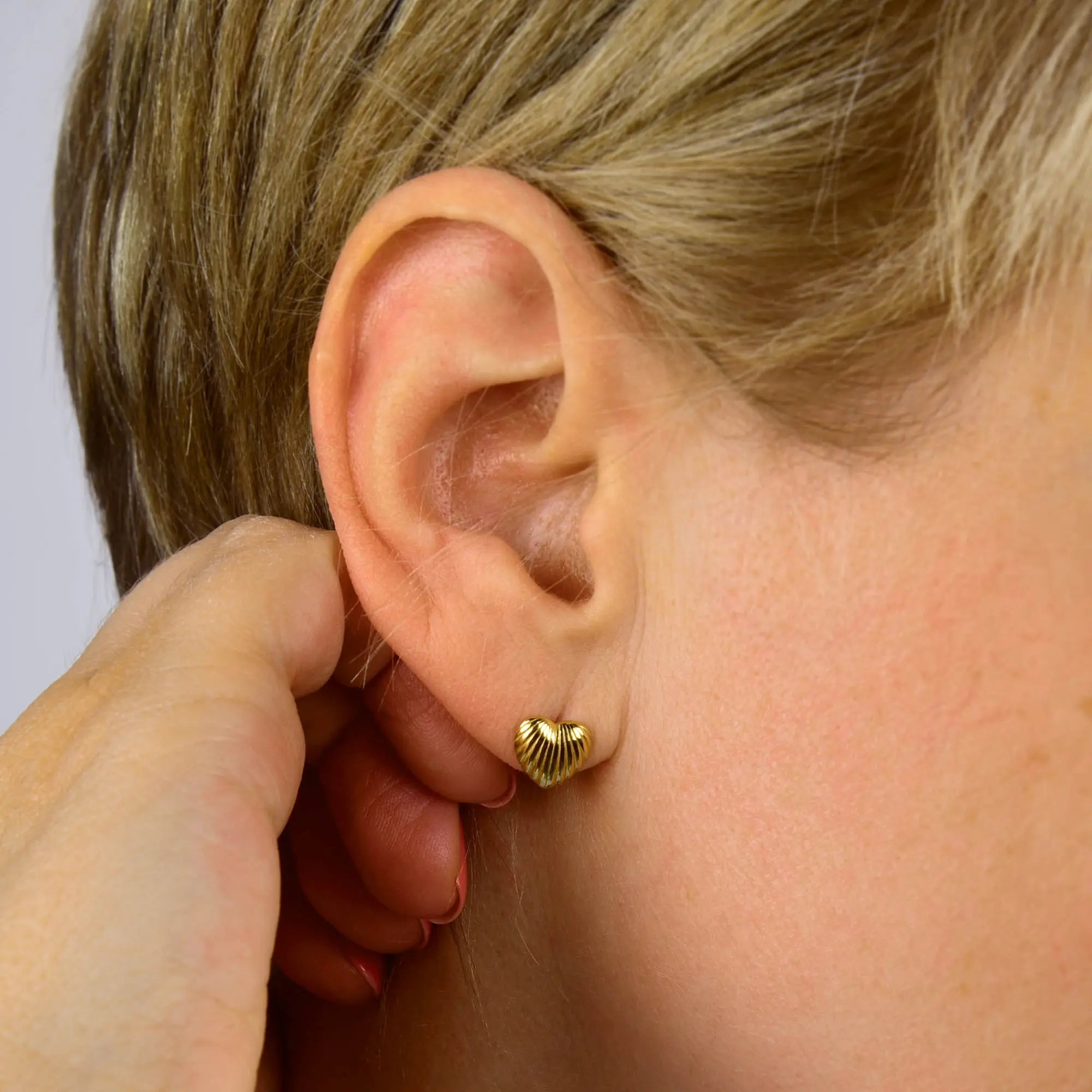 A close-up of a person’s ear wearing a small, gold Heart Beam Stud Earring in the shape of a heart. The person has short, light brown hair, and a hand is gently touching the ear.