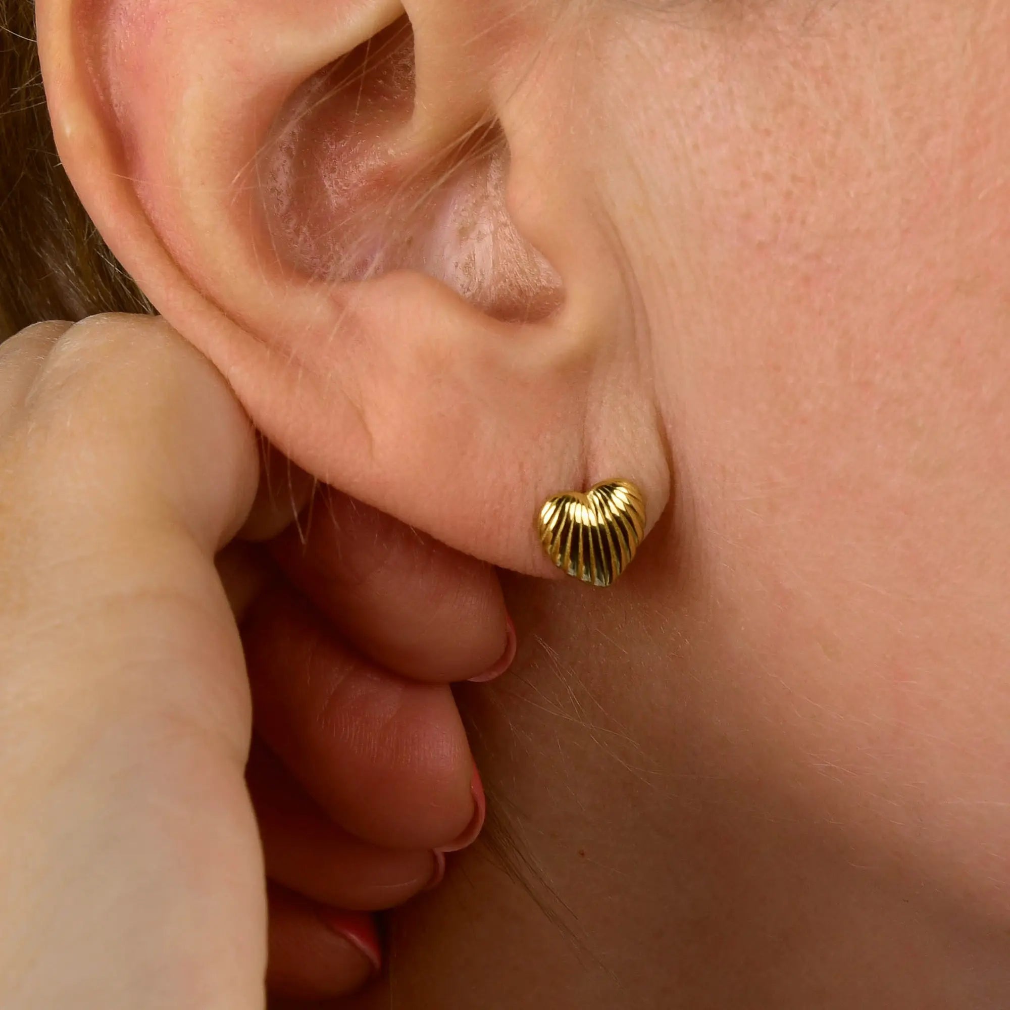 Close-up of a person’s ear adorned with a small, gold, heart-shaped stud earring featuring a ribbed texture, reminiscent of the Heart Beam Stud Earrings. The person’s hand is near their face, with part of their fingers visible.