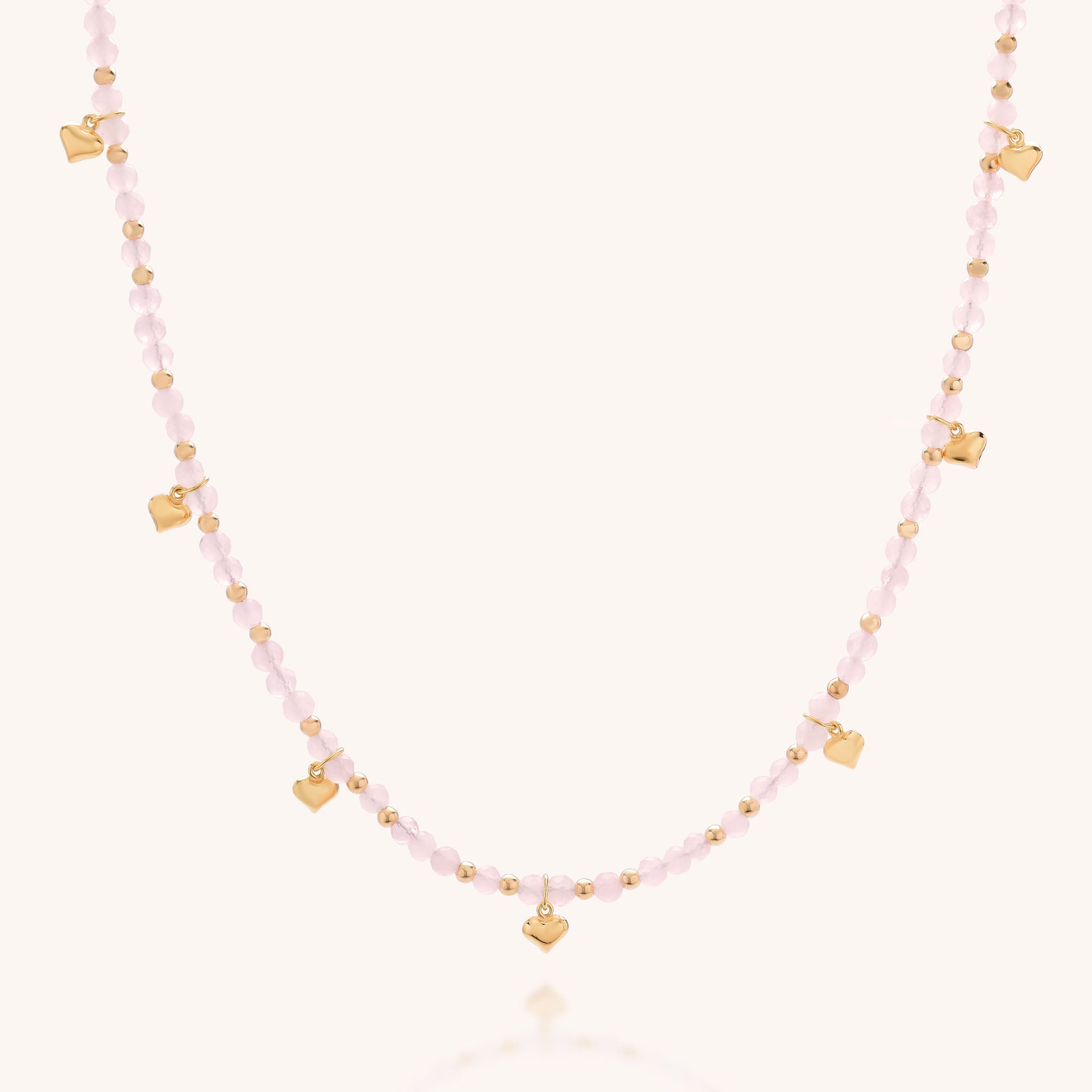 The Rose Hearts Necklace features light pink beads, small gold rounds, and tiny gold heart charms evenly spaced along the chain, shown on a plain light background.