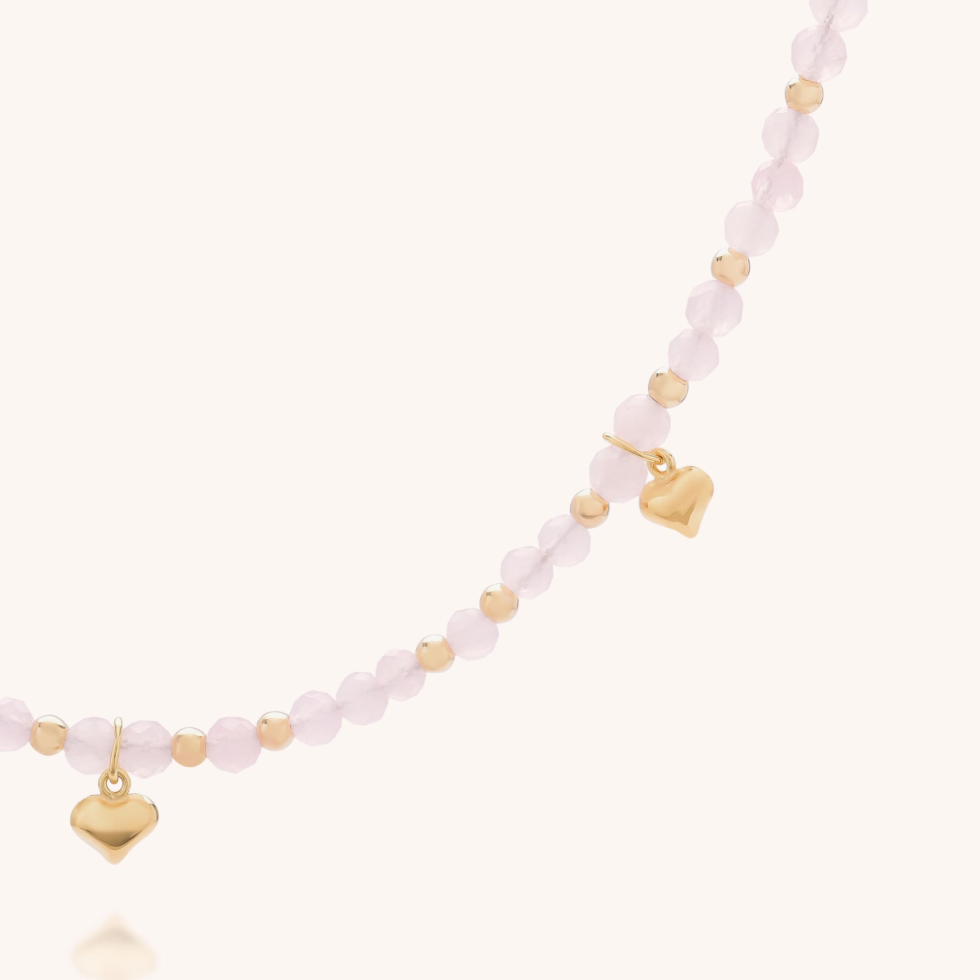 The Rose Hearts Necklace features small round pale pink beads, gold spacers, and two gold heart charms. Set against a soft light cream background, this delicate necklace adds a touch of romance to any look.